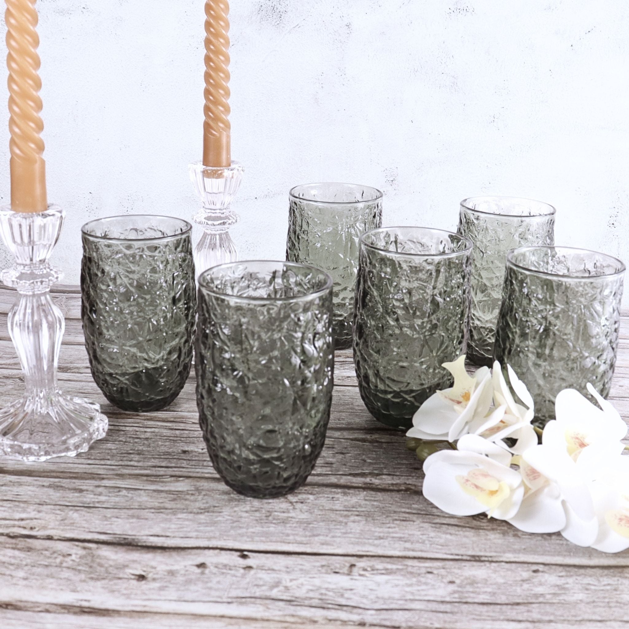 Set of textured gray glasses on a wooden surface with white flowers and candle holders.