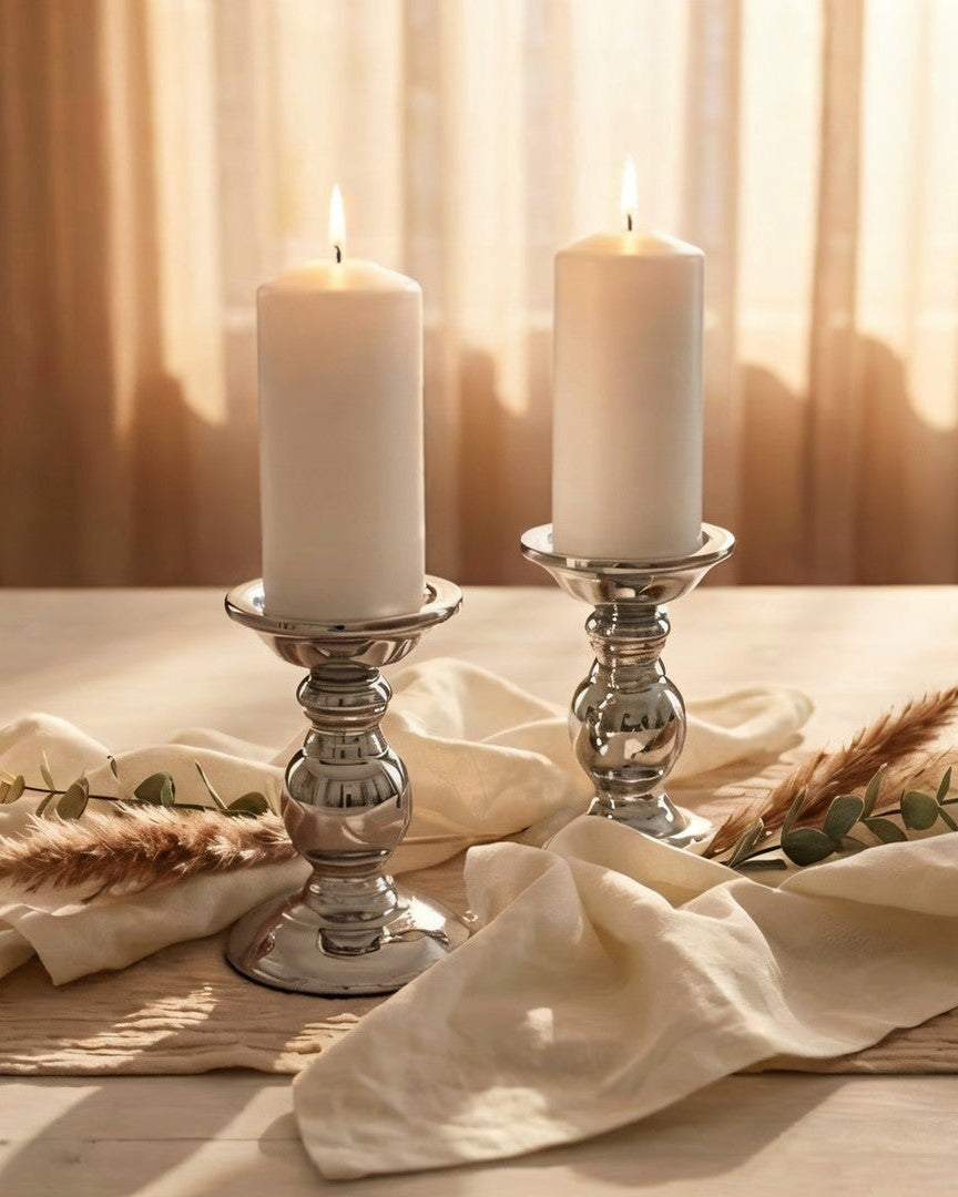 Two white candles in silver holders on a wooden table with a soft, warm light.
