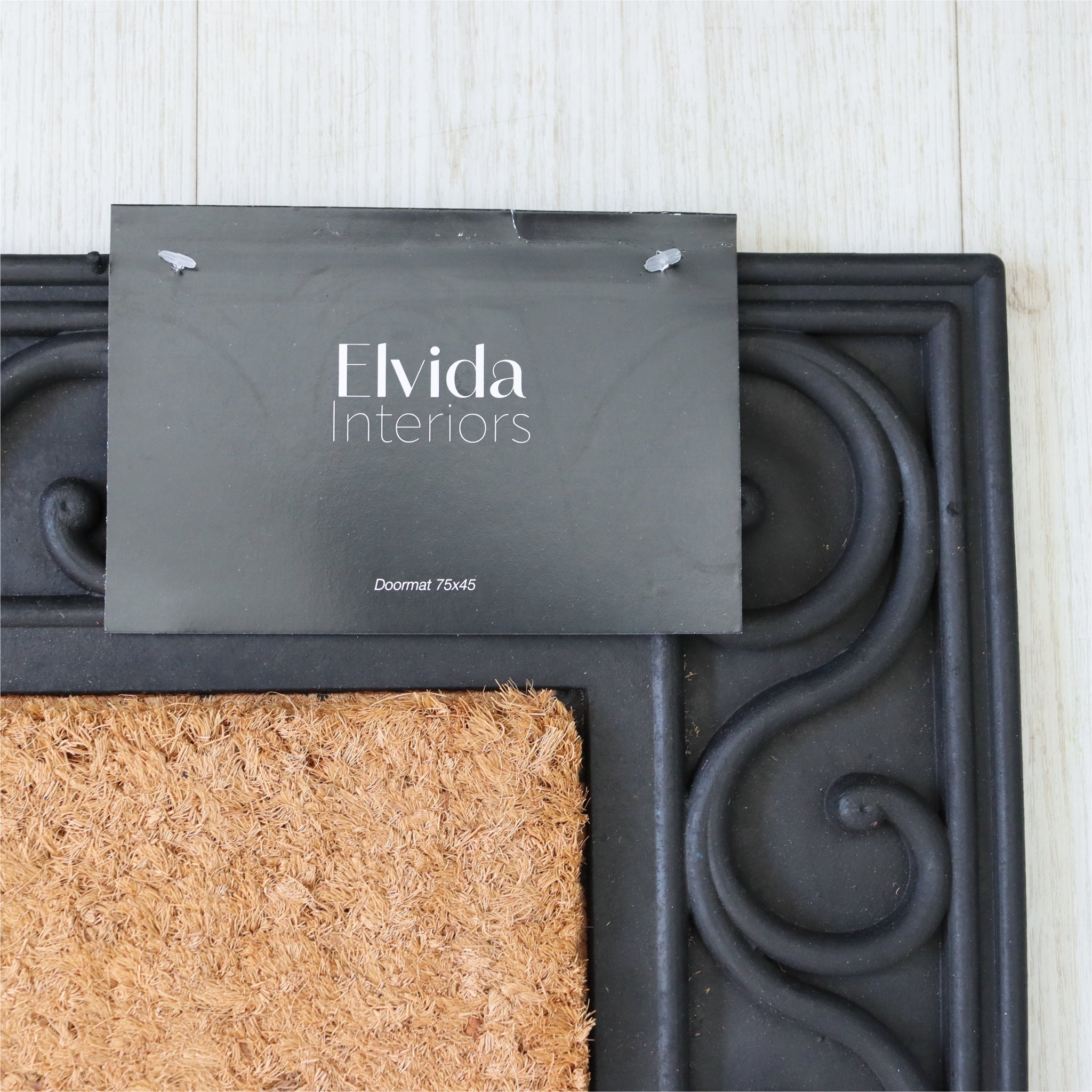 Doormat with decorative border and 'Elvida Interiors' sign on a white wall background