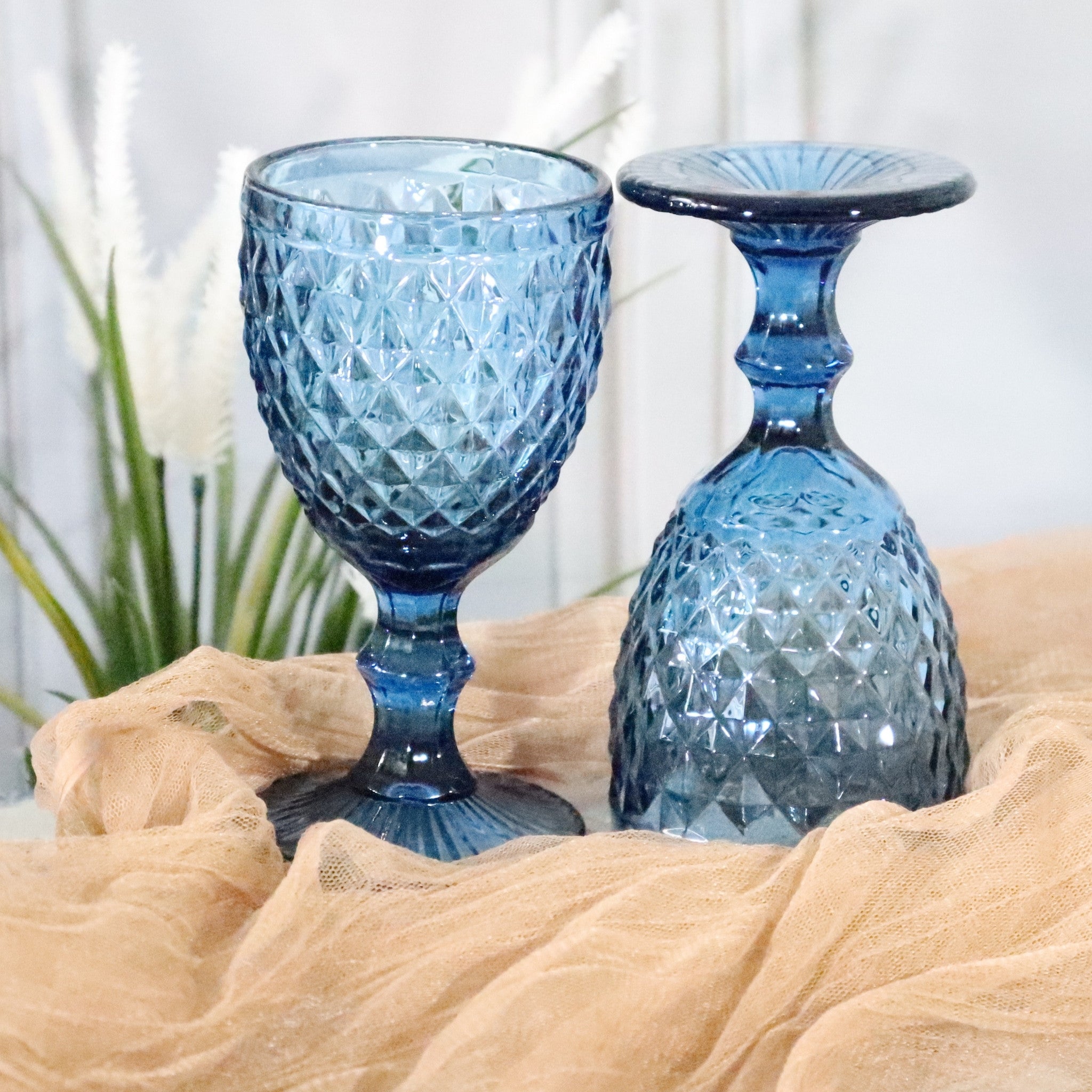 Two blue textured glass goblets on a beige surface with a blurred plant background