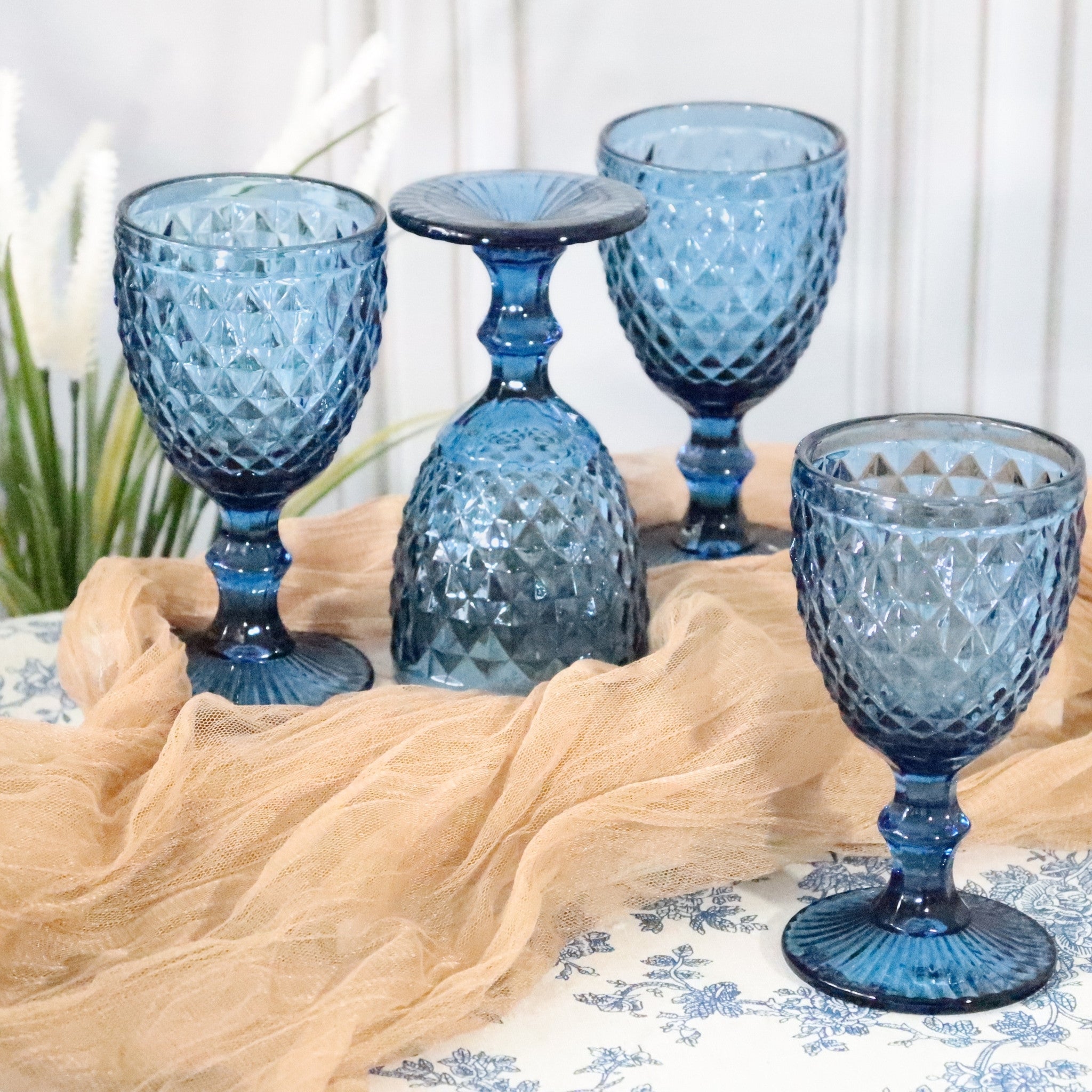 Set of blue textured glass goblets on a patterned tablecloth with a neutral fabric background.