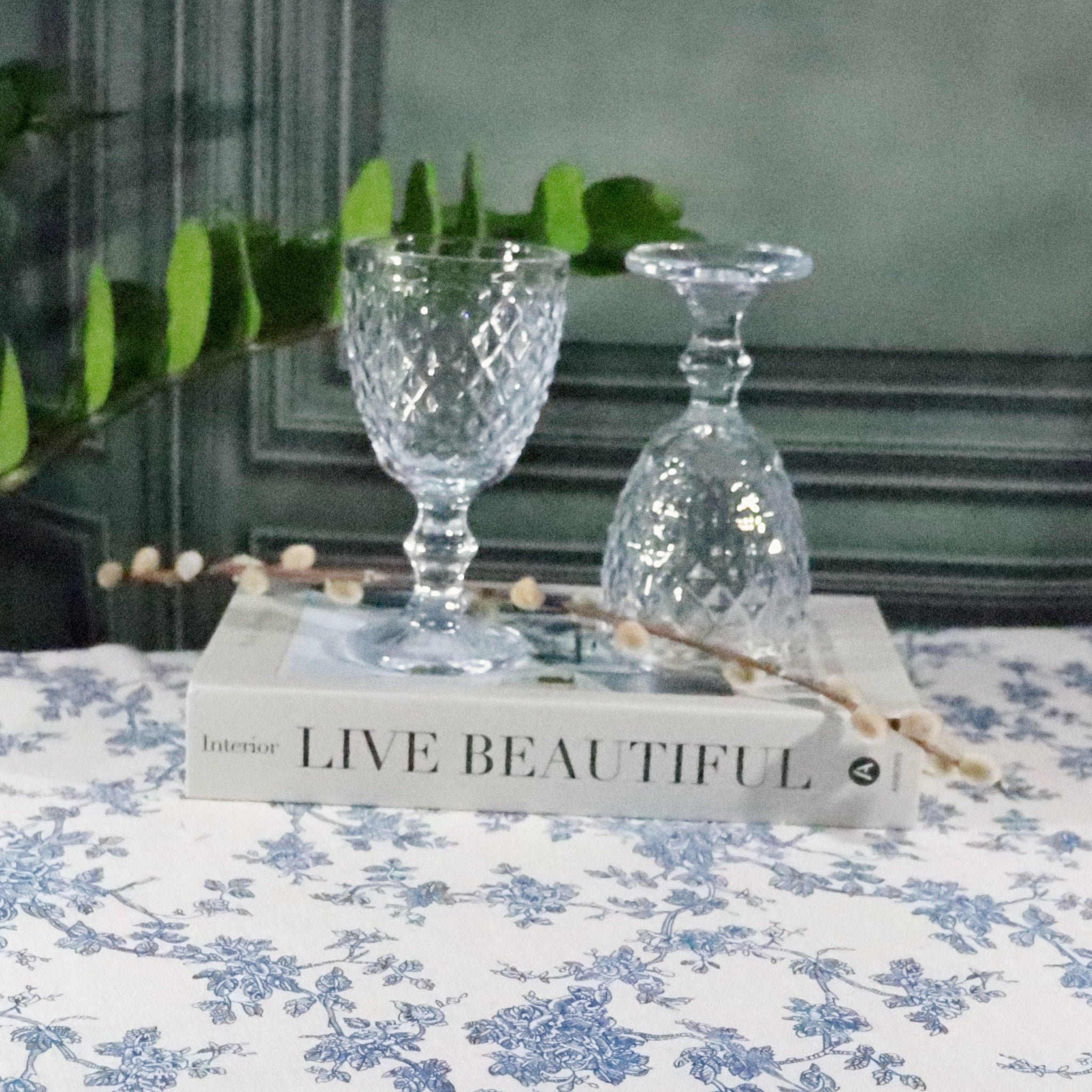 Two clear glass goblets on a book titled 'Live Beautiful' with a floral tablecloth and plant in the background.