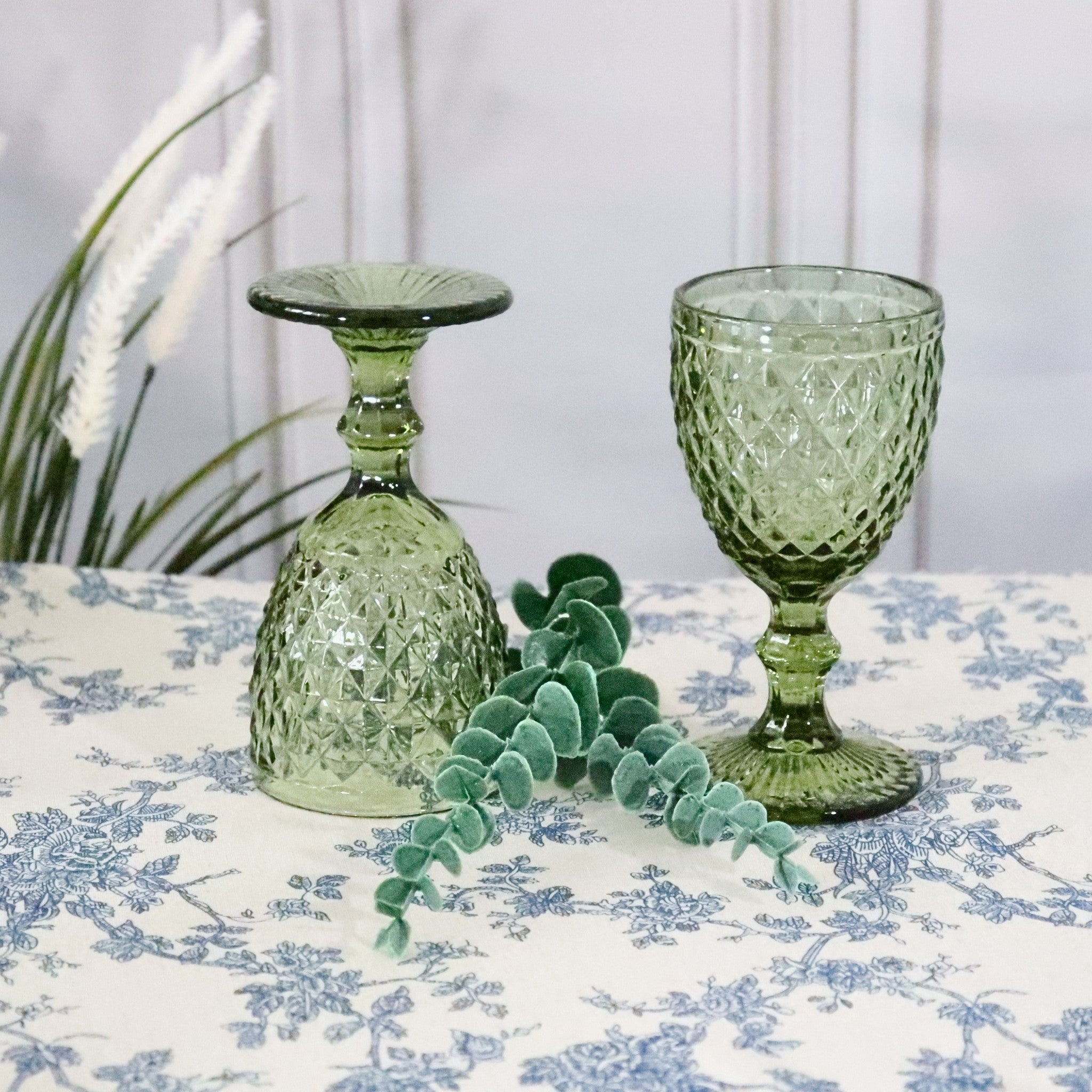 Two green textured glass goblets on a floral tablecloth with a plant in the background.