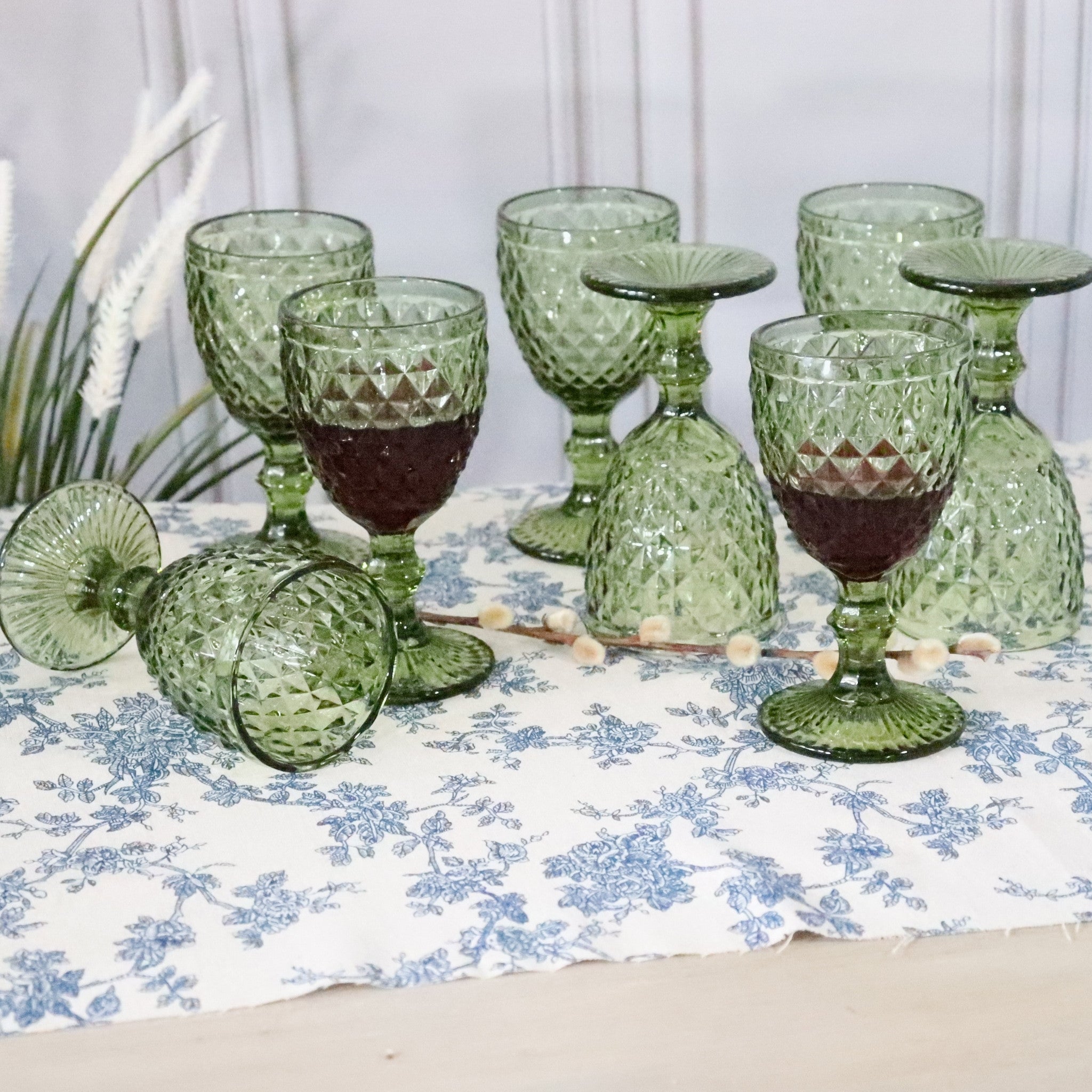 Set of green glass goblets on a floral tablecloth with a white curtain background