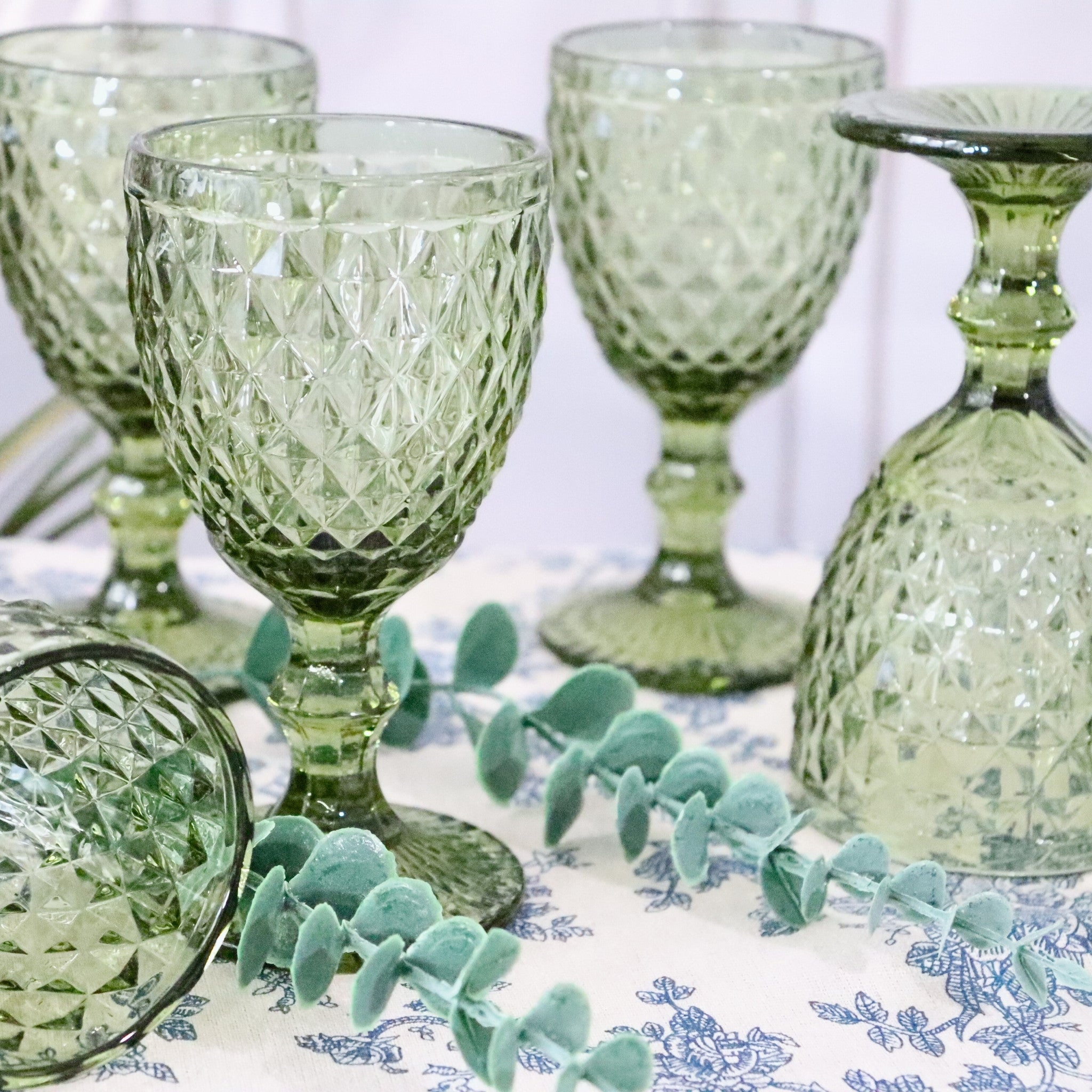 Set of green glass goblets on a decorative tablecloth with eucalyptus leaves.