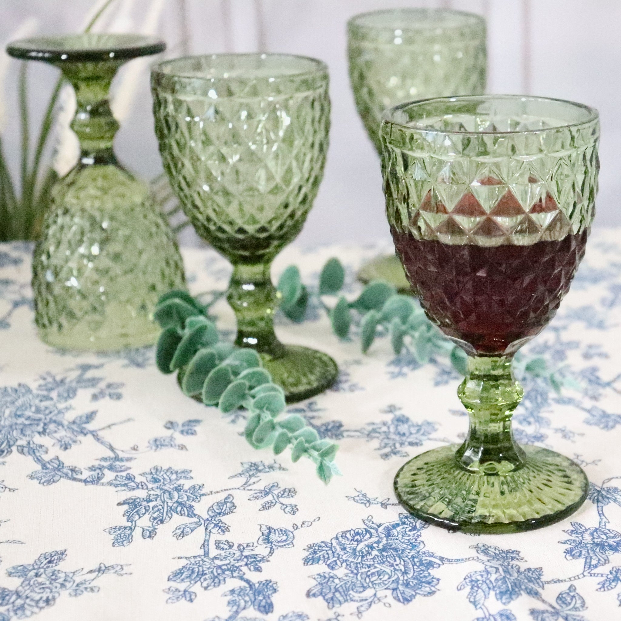 Green glass goblets on a floral tablecloth with one containing a dark red liquid.