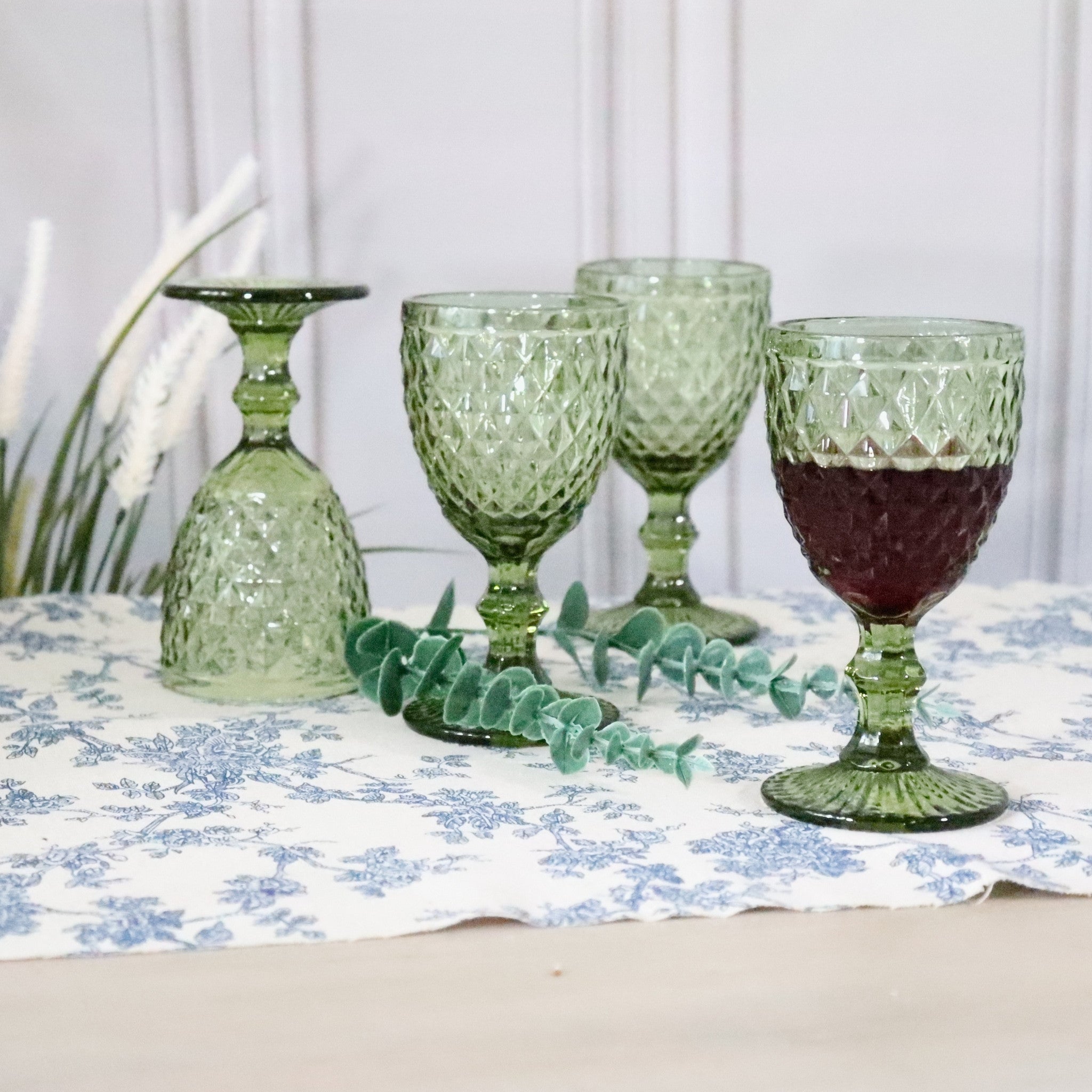 Green glassware on a table with a floral tablecloth and eucalyptus leaves.