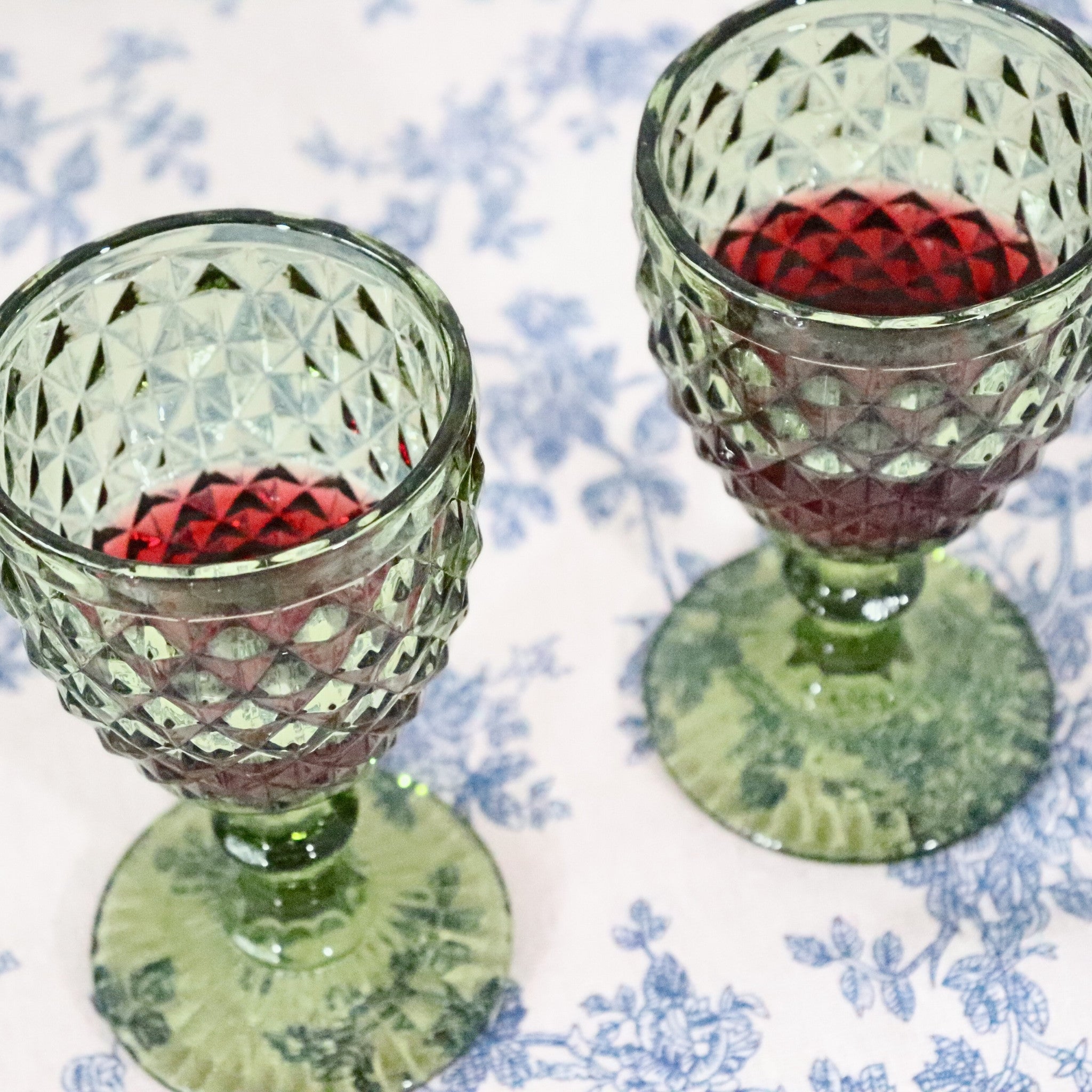 Two green patterned glasses with red liquid on a floral tablecloth