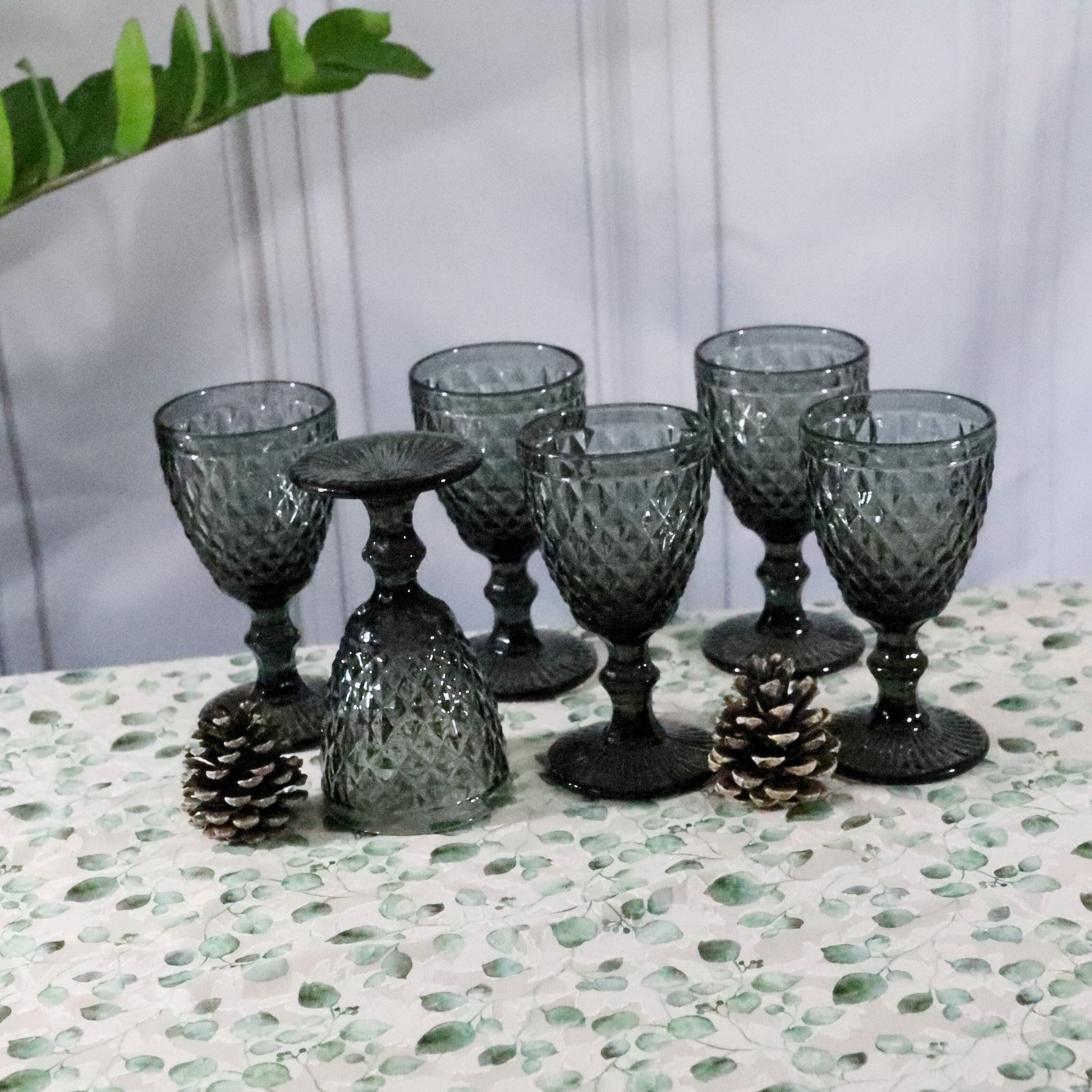 Set of six dark gray textured glass goblets on a patterned surface with decorative pinecones.