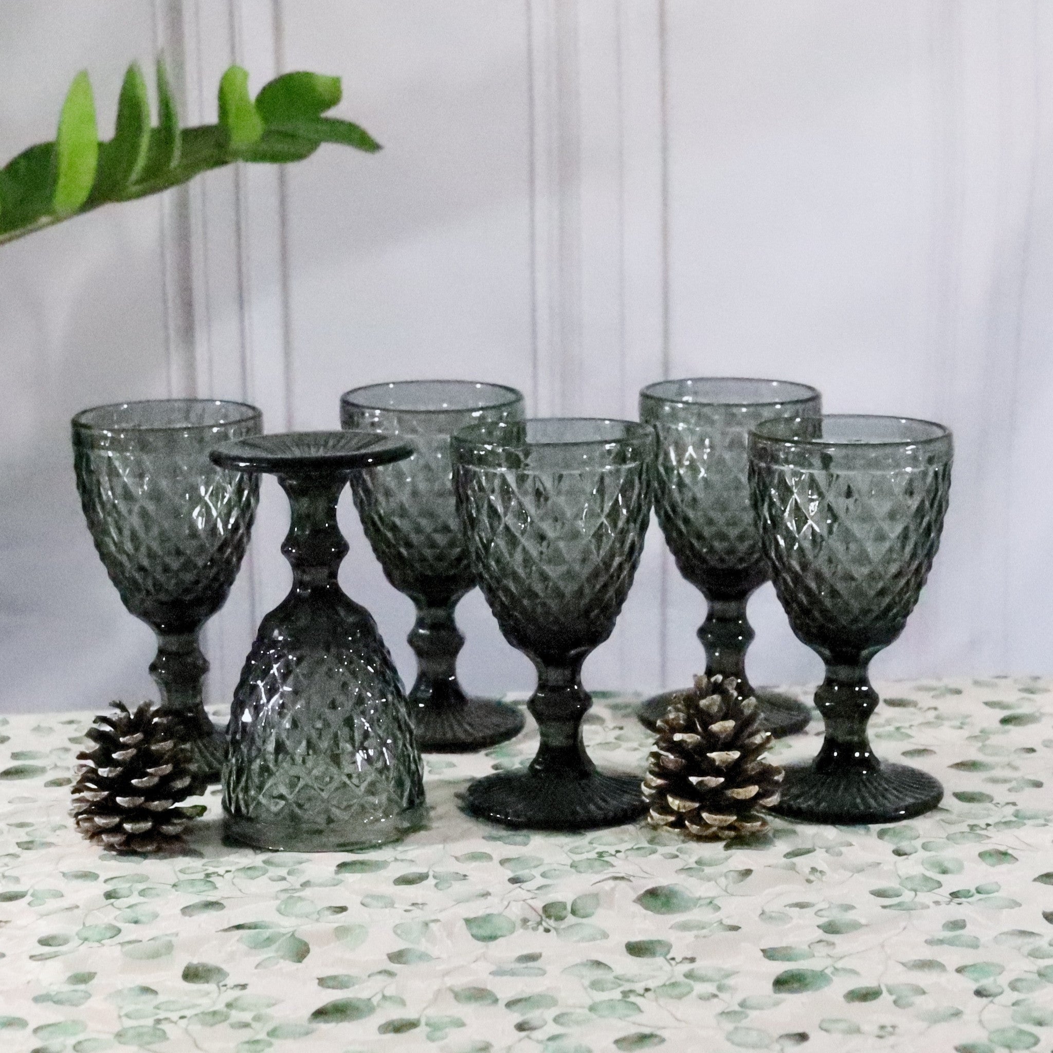 Set of six gray glass goblets on a textured surface with decorative pinecones.