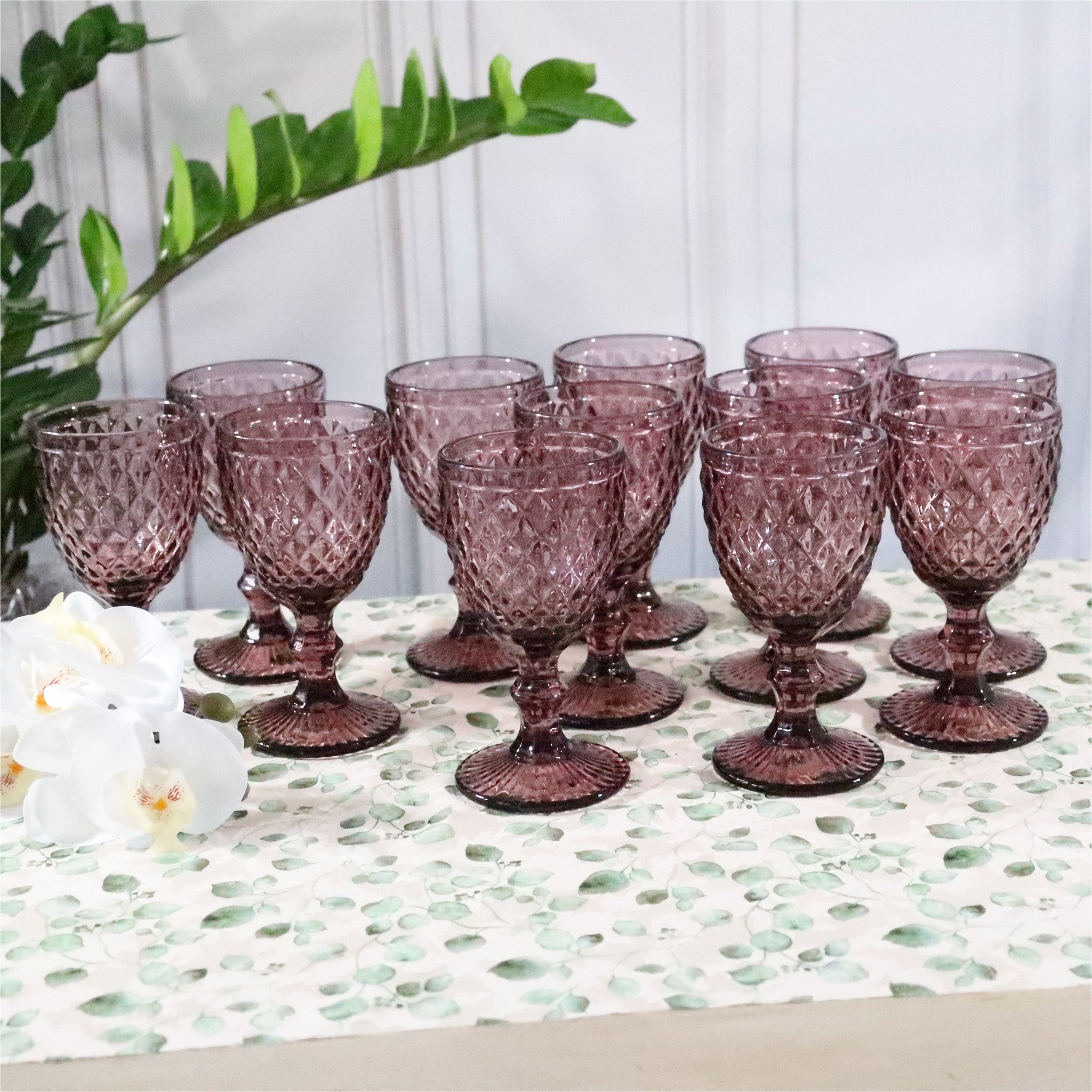 Set of purple glass goblets on a decorative tablecloth with a plant and flowers in the background.