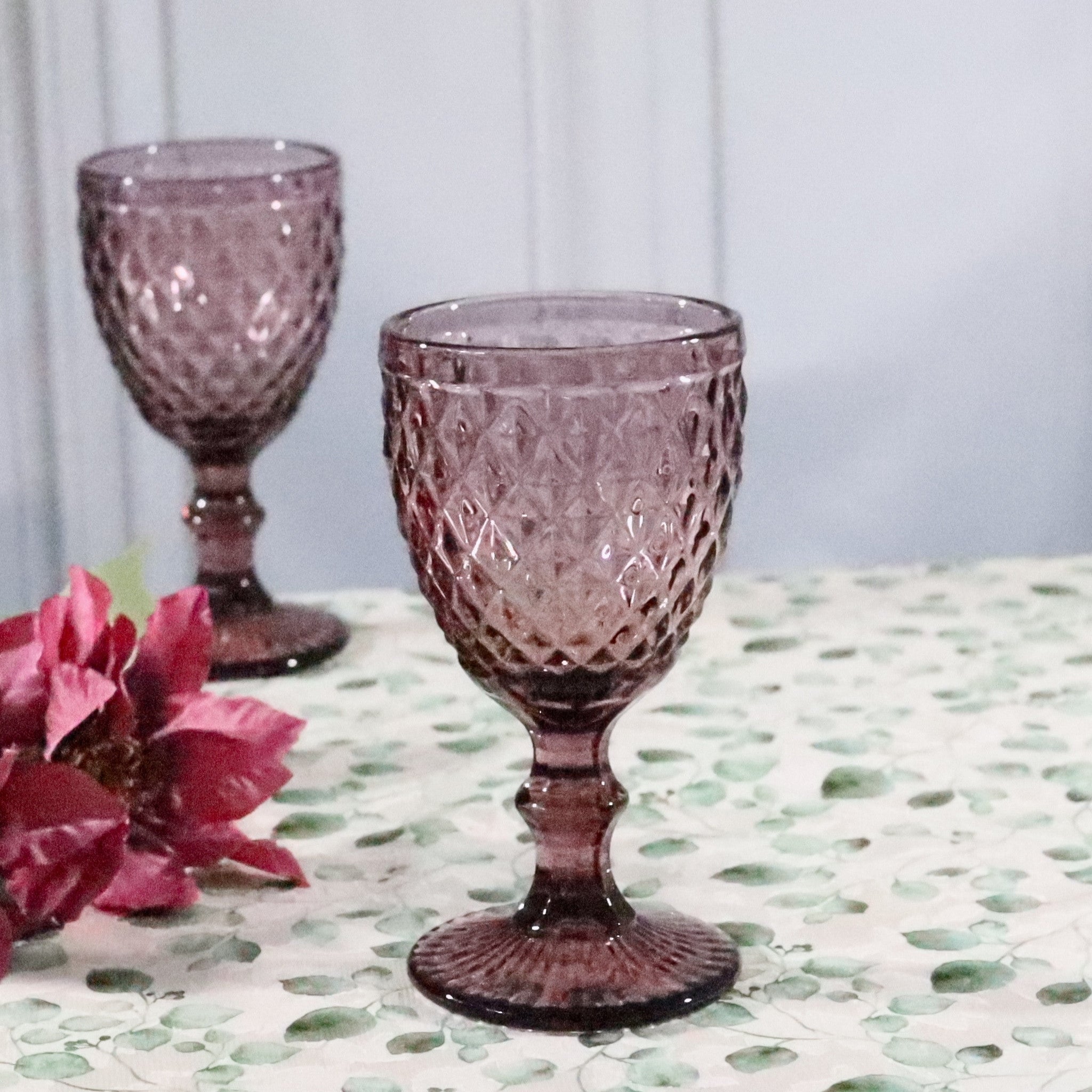 Two purple textured glass goblets on a patterned surface with pink flowers.