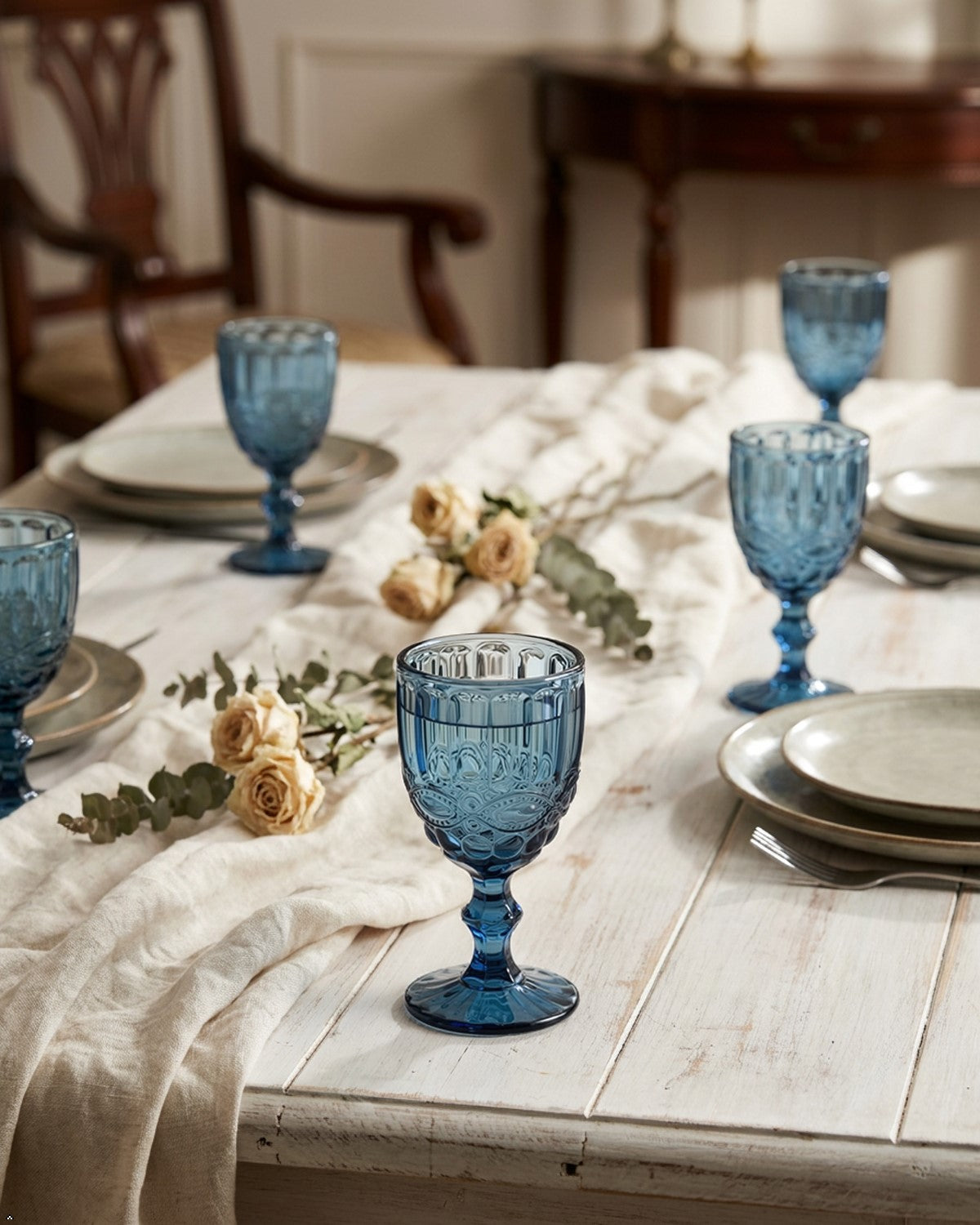 Dining table set with blue glass goblets and white plates on a wooden table.