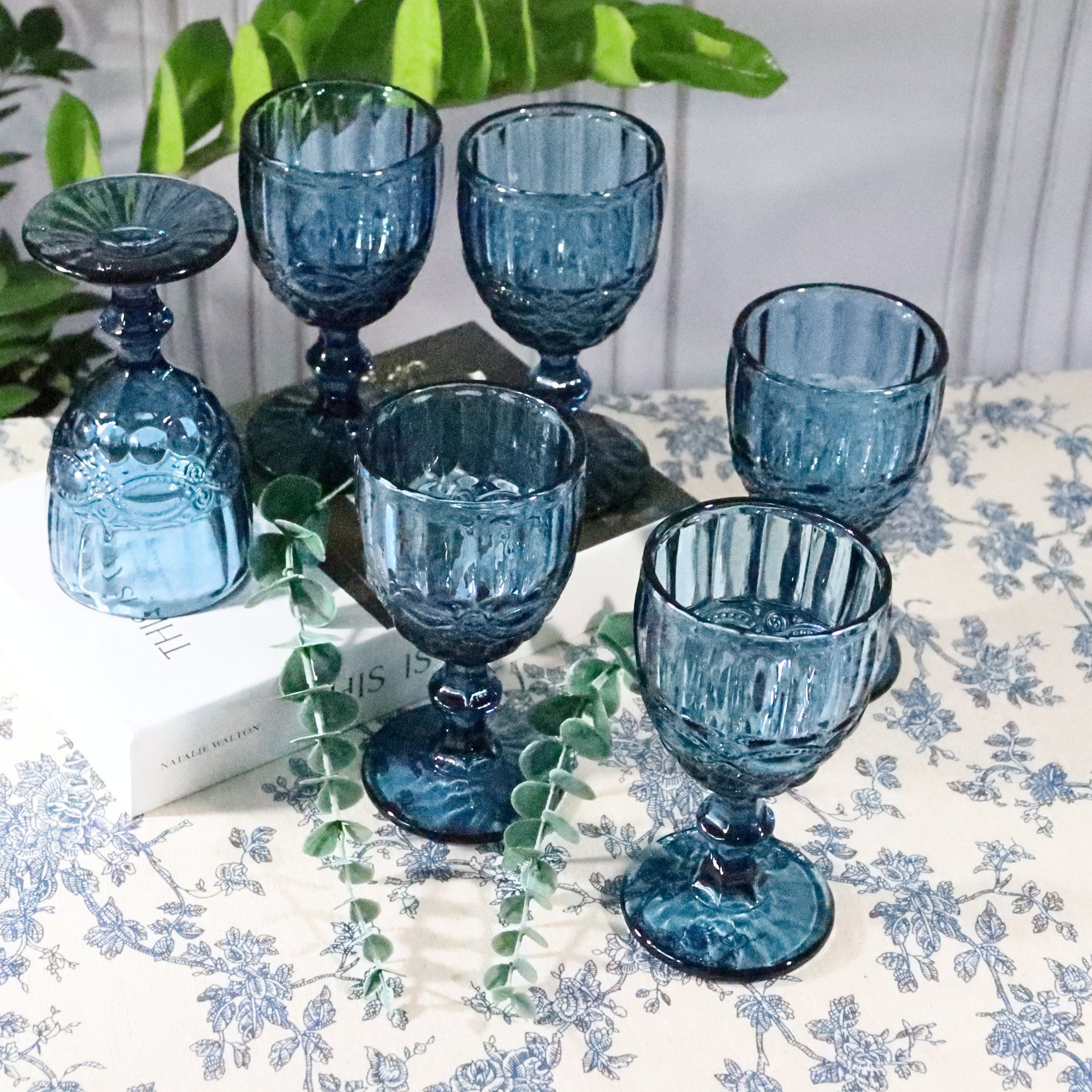 Set of six blue glass goblets on a floral tablecloth with green leaves in the background