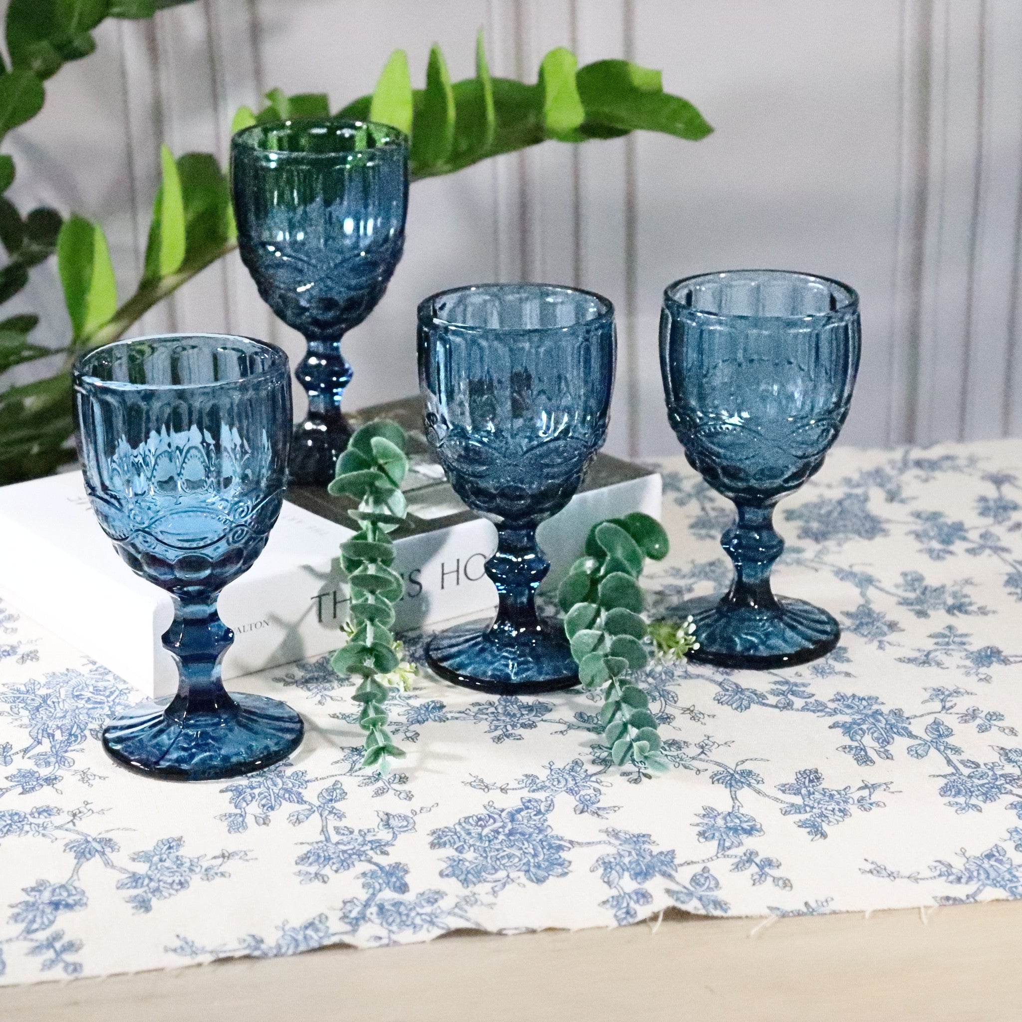 Set of blue glass goblets on a floral tablecloth with greenery.
