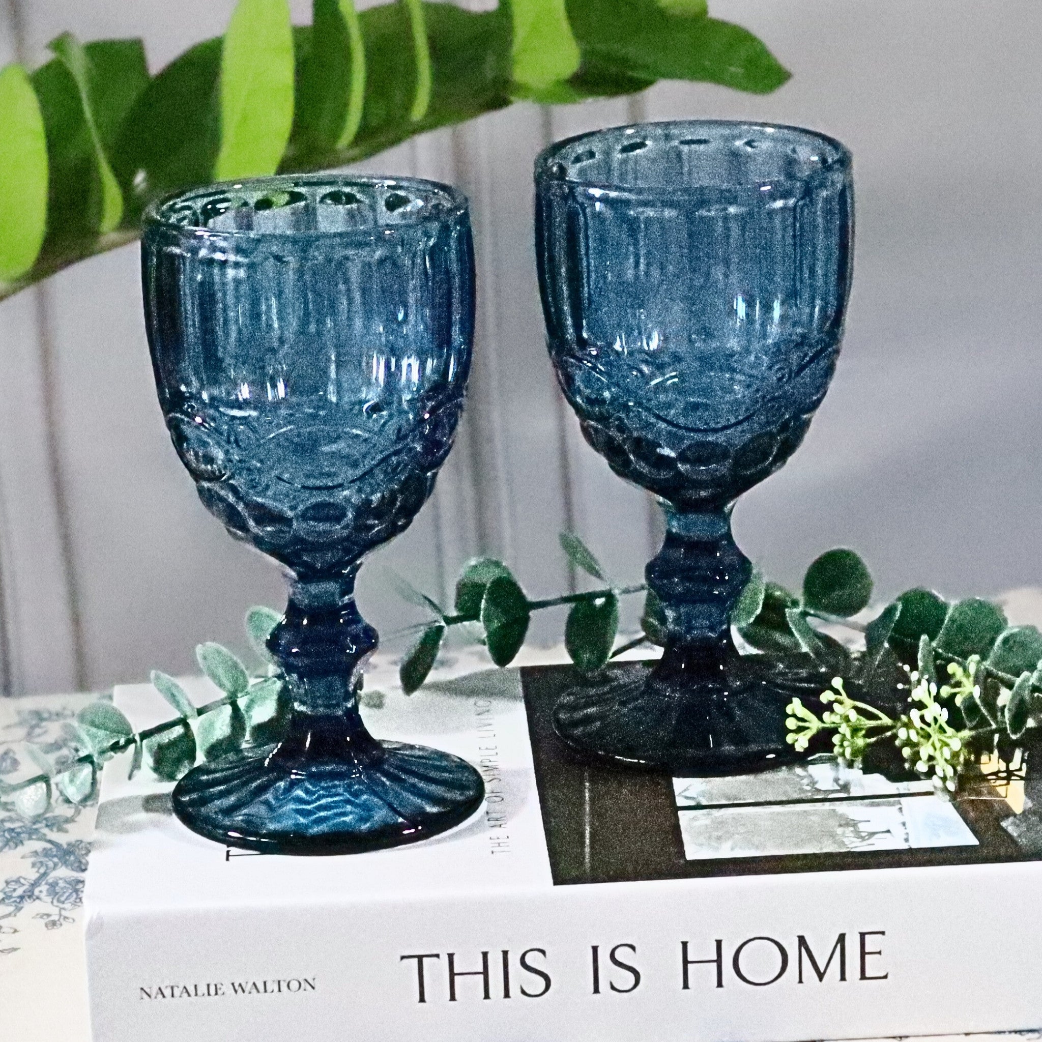 Two blue textured glass goblets on a surface with greenery and a book titled 'This is Home' in the background.