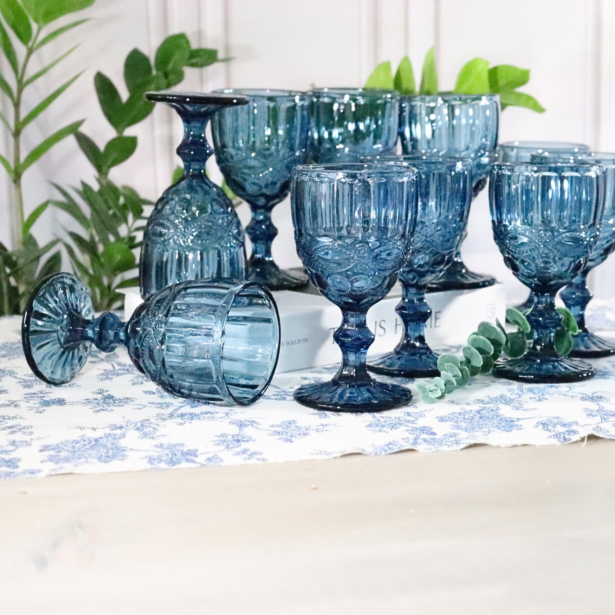 Set of blue glass goblets on a table with a white and blue patterned tablecloth and green plants in the background.
