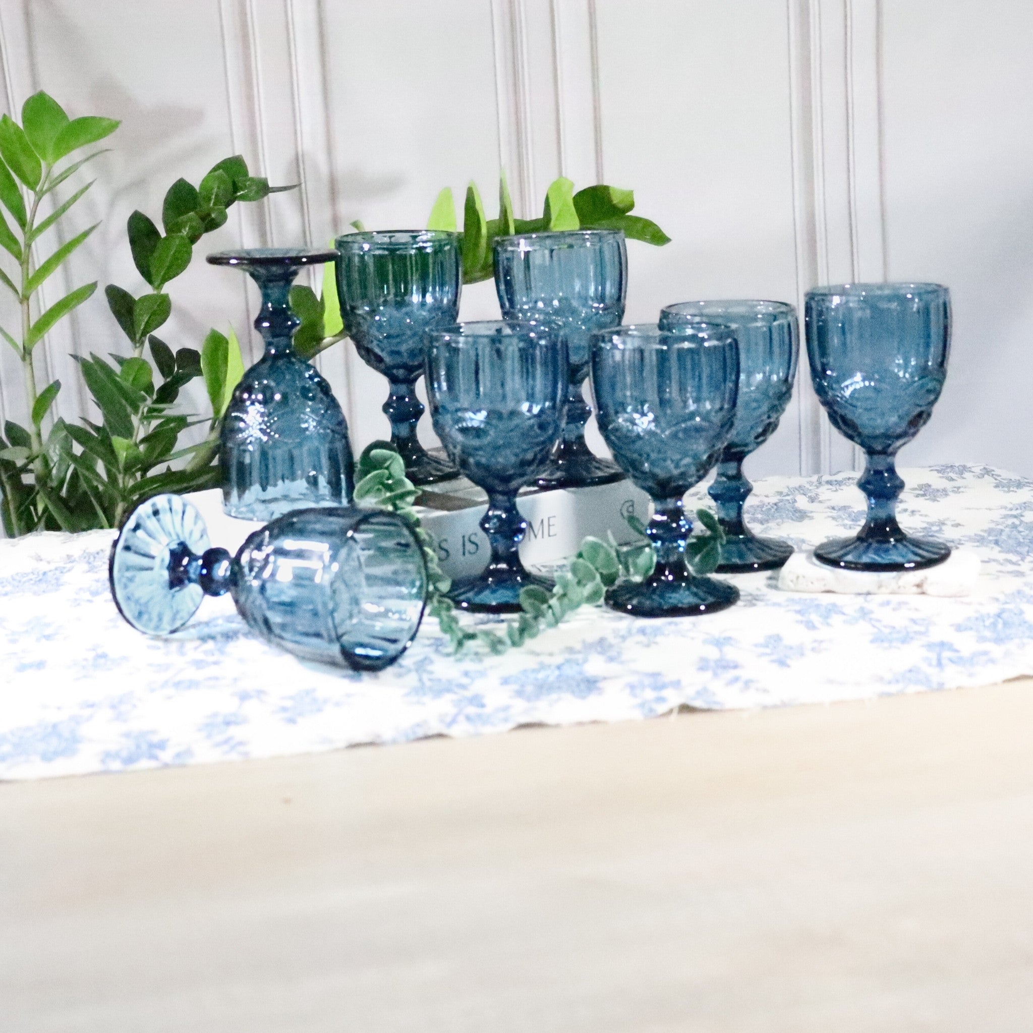 Set of blue glassware on a table with a white cushion and green plants in the background