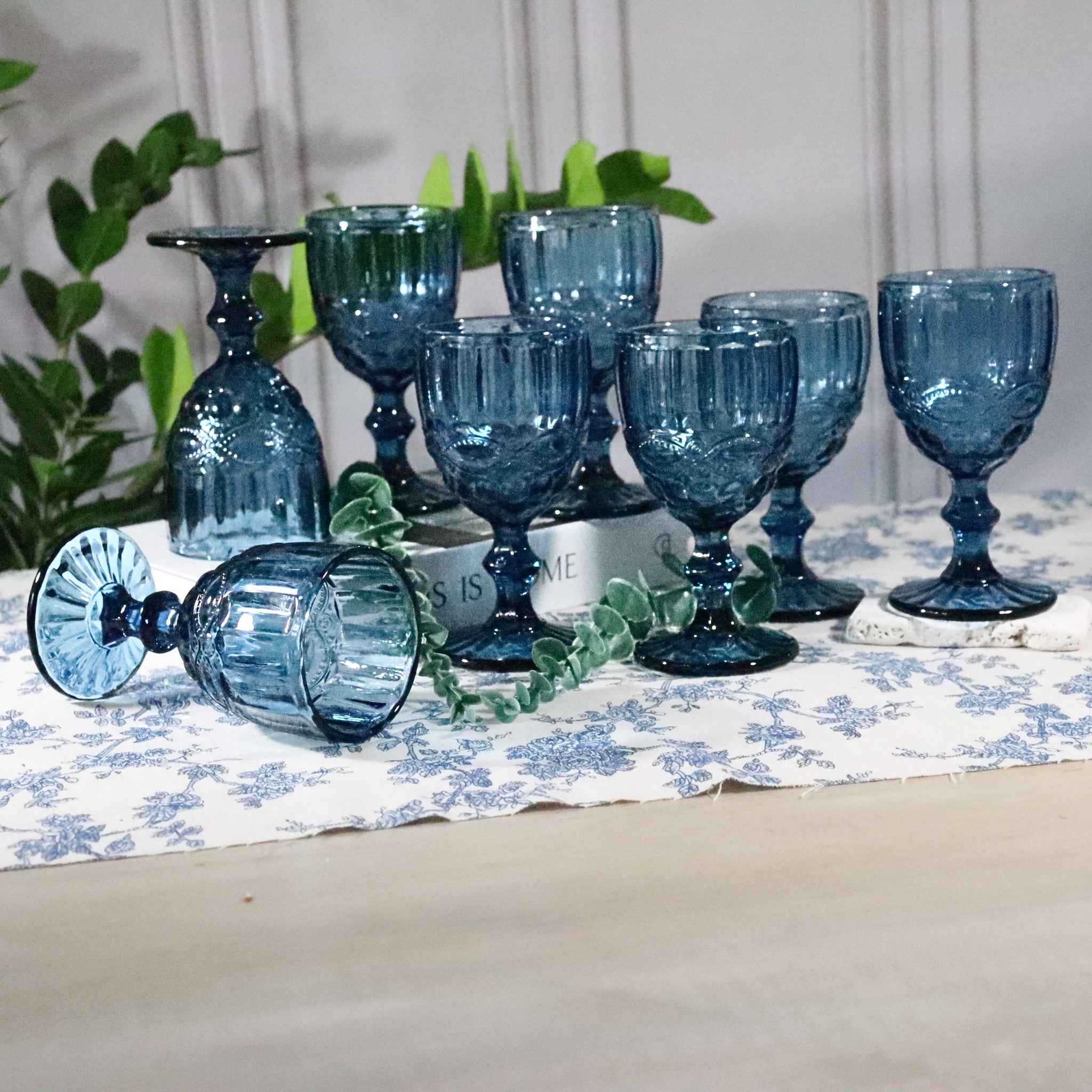 Set of blue glassware on a table with a floral tablecloth and plants in the background.