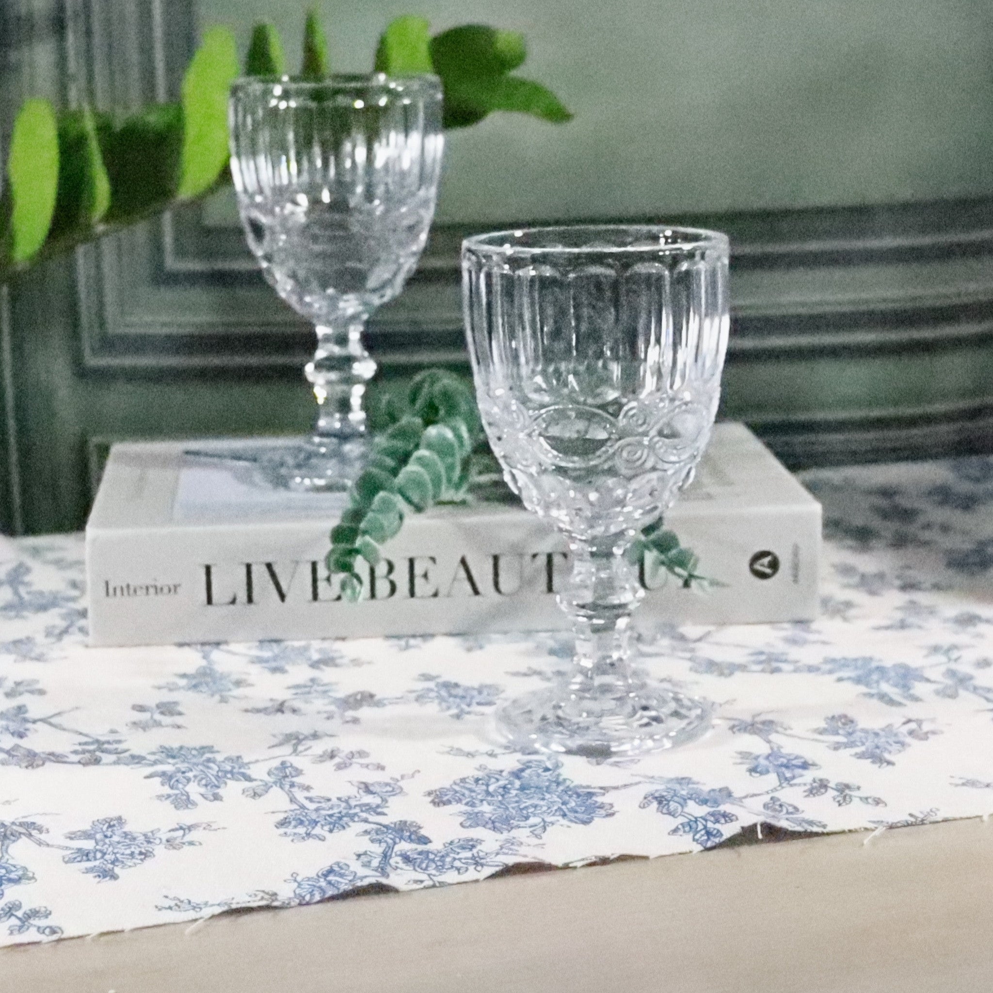 Clear glass goblets on a book with a floral tablecloth and plant in the background