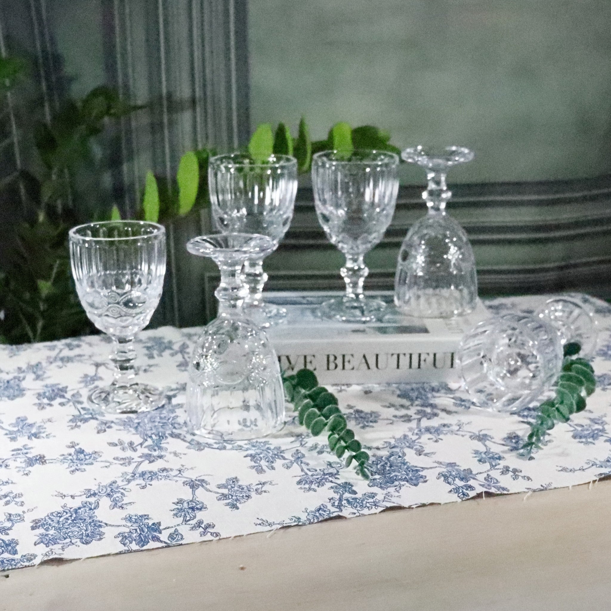 Set of clear glass goblets on a floral tablecloth with a plant in the background