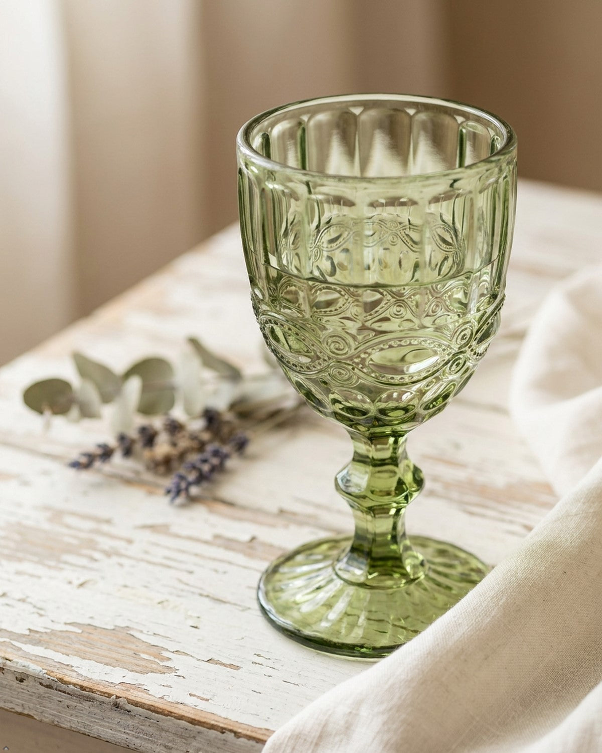 Green glass goblet with intricate patterns on a wooden surface with a white cloth and lavender flowers.