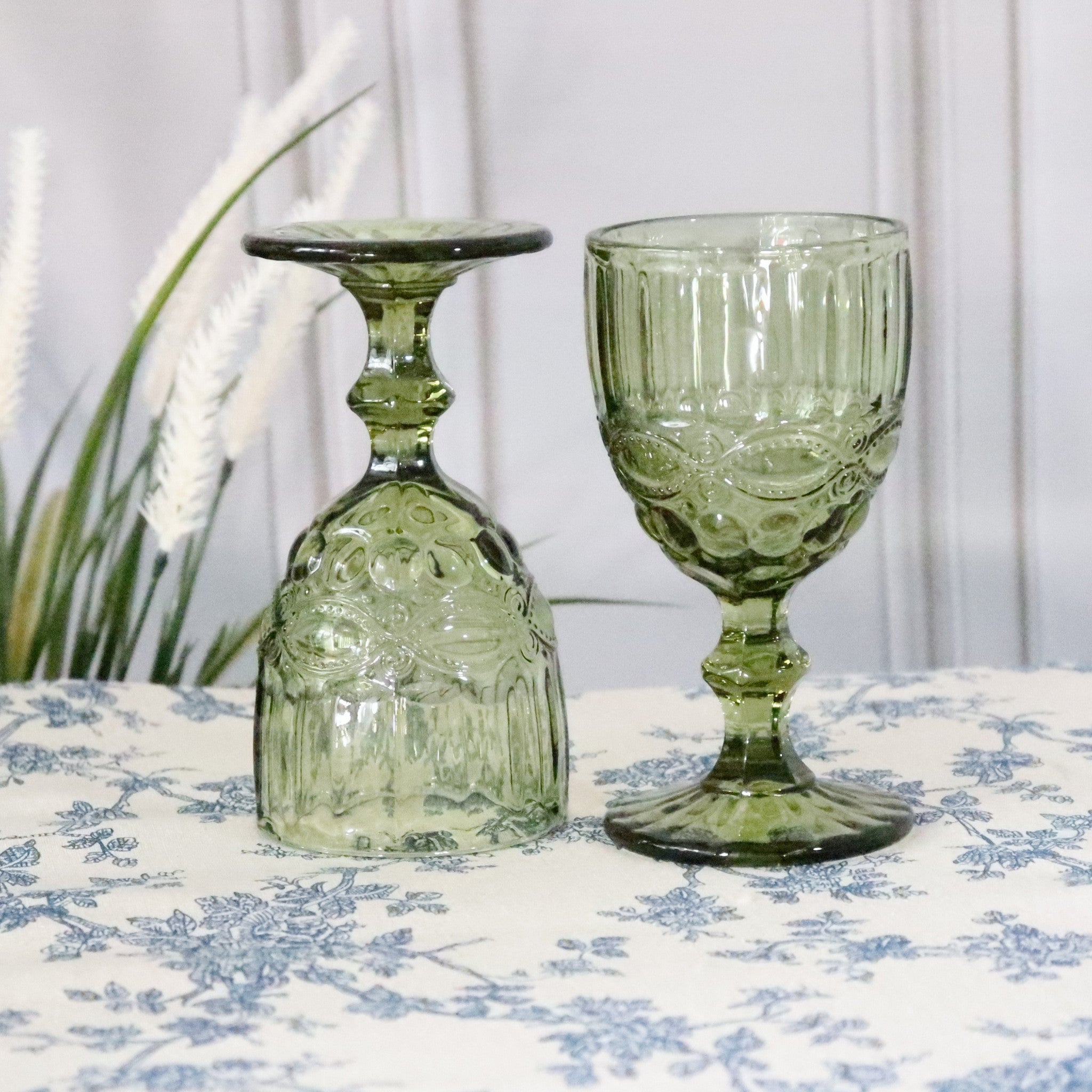 Two green glass goblets on a floral tablecloth with a white curtain background