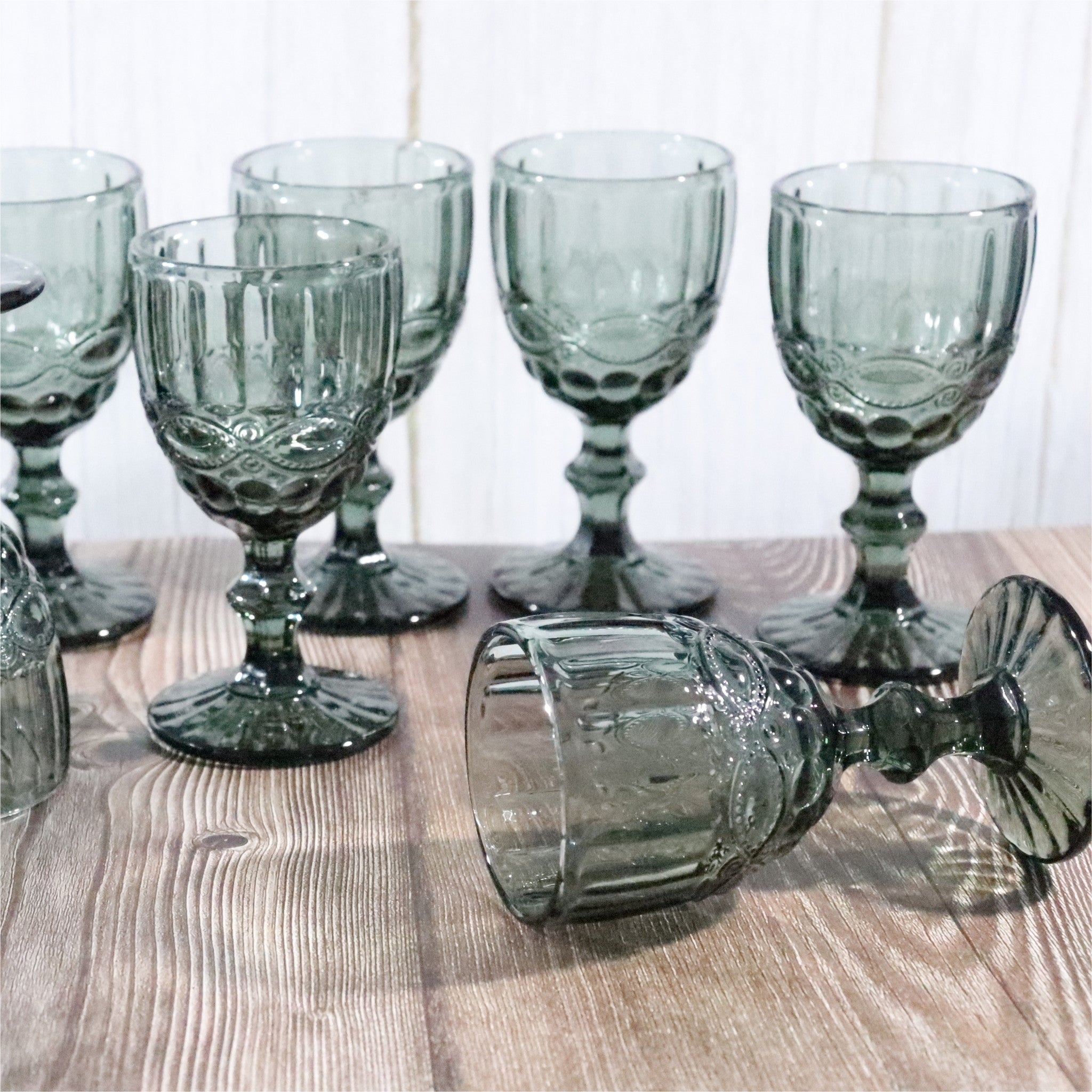 Set of six grey glass goblets on a wooden surface with a white tiled wall in the background.
