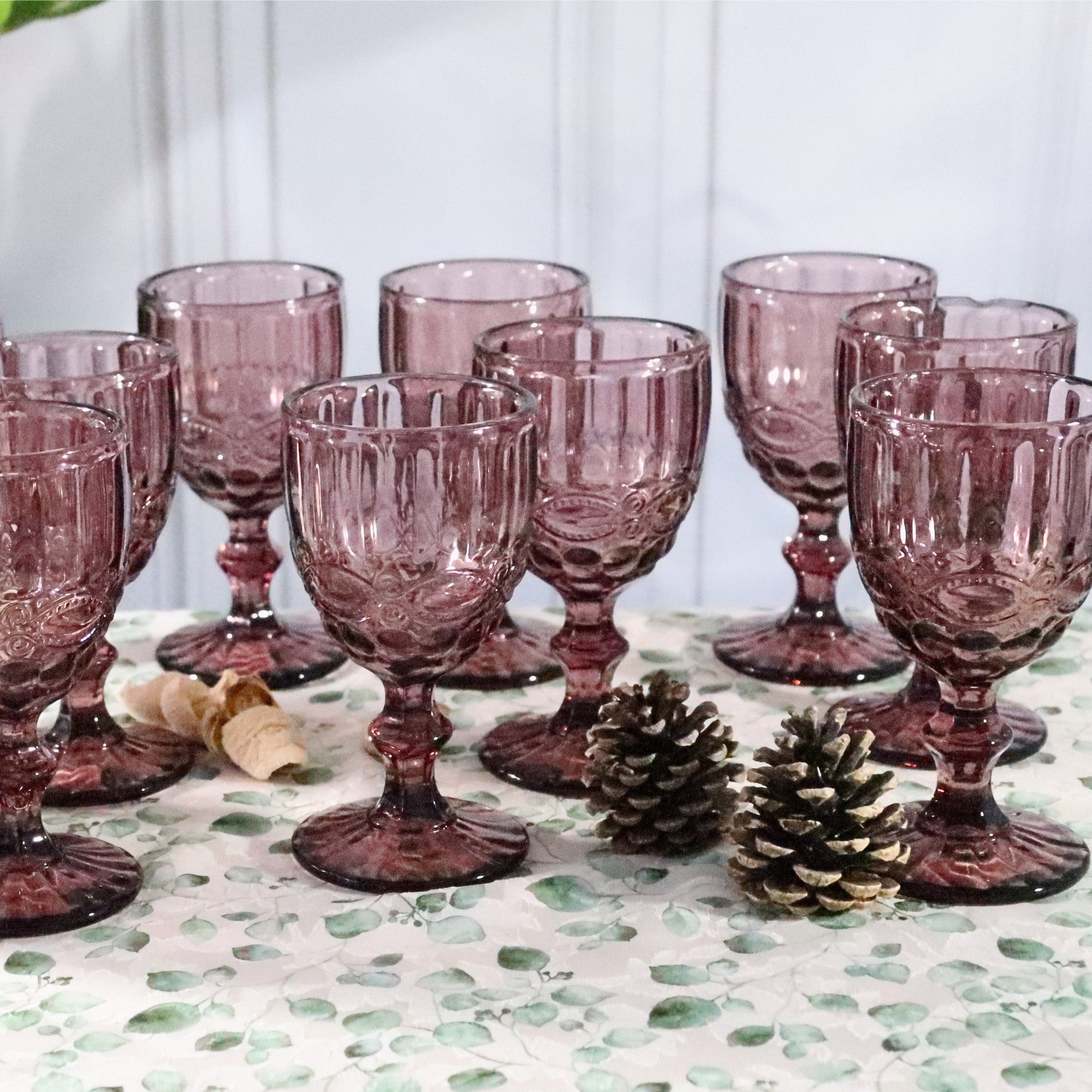 Set of purple glass goblets on a decorative tablecloth with pine cones.