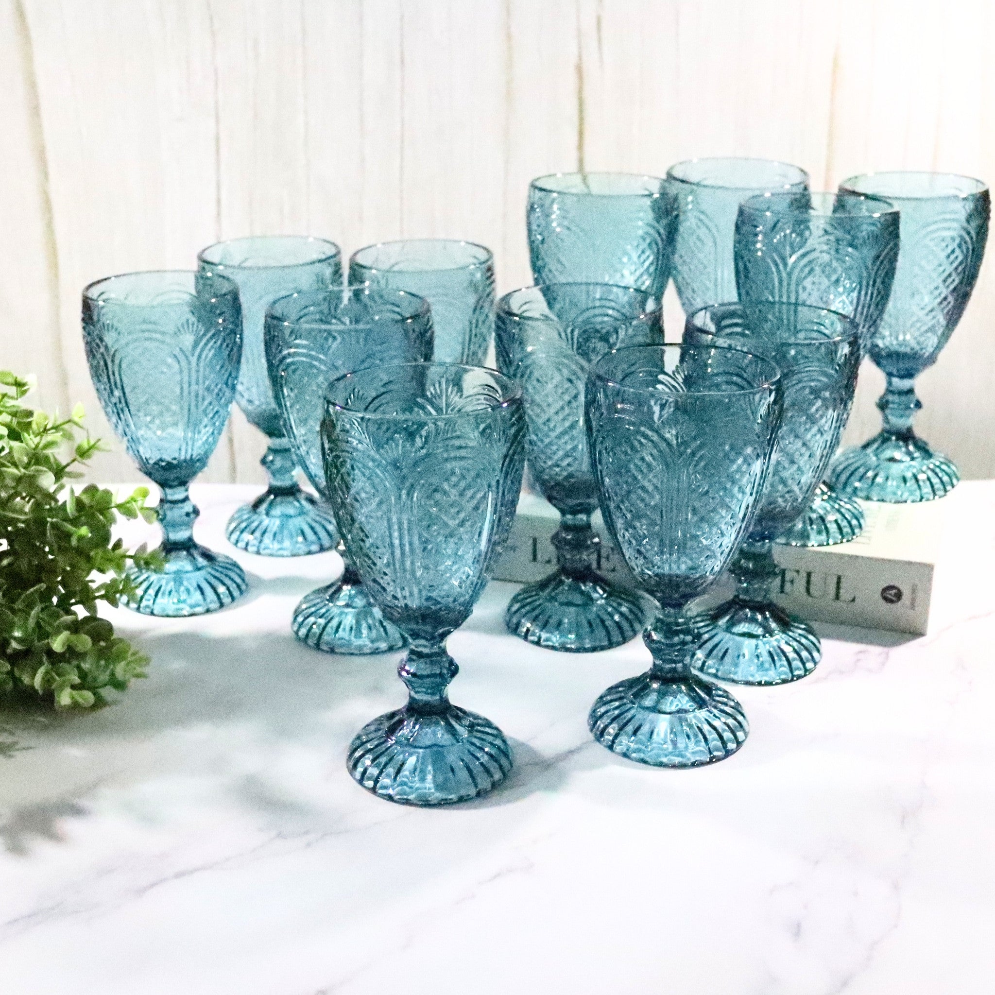 Set of blue glass goblets on a white surface with a plant and book in the background.