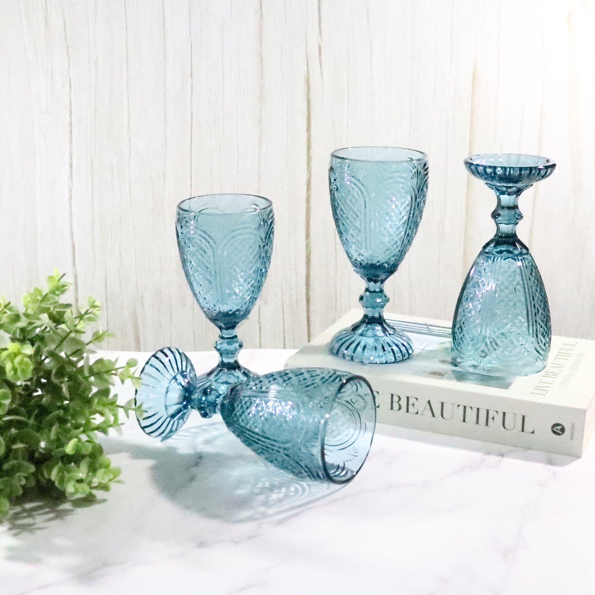 Set of blue textured glass goblets on a white surface with a plant and book in the background.