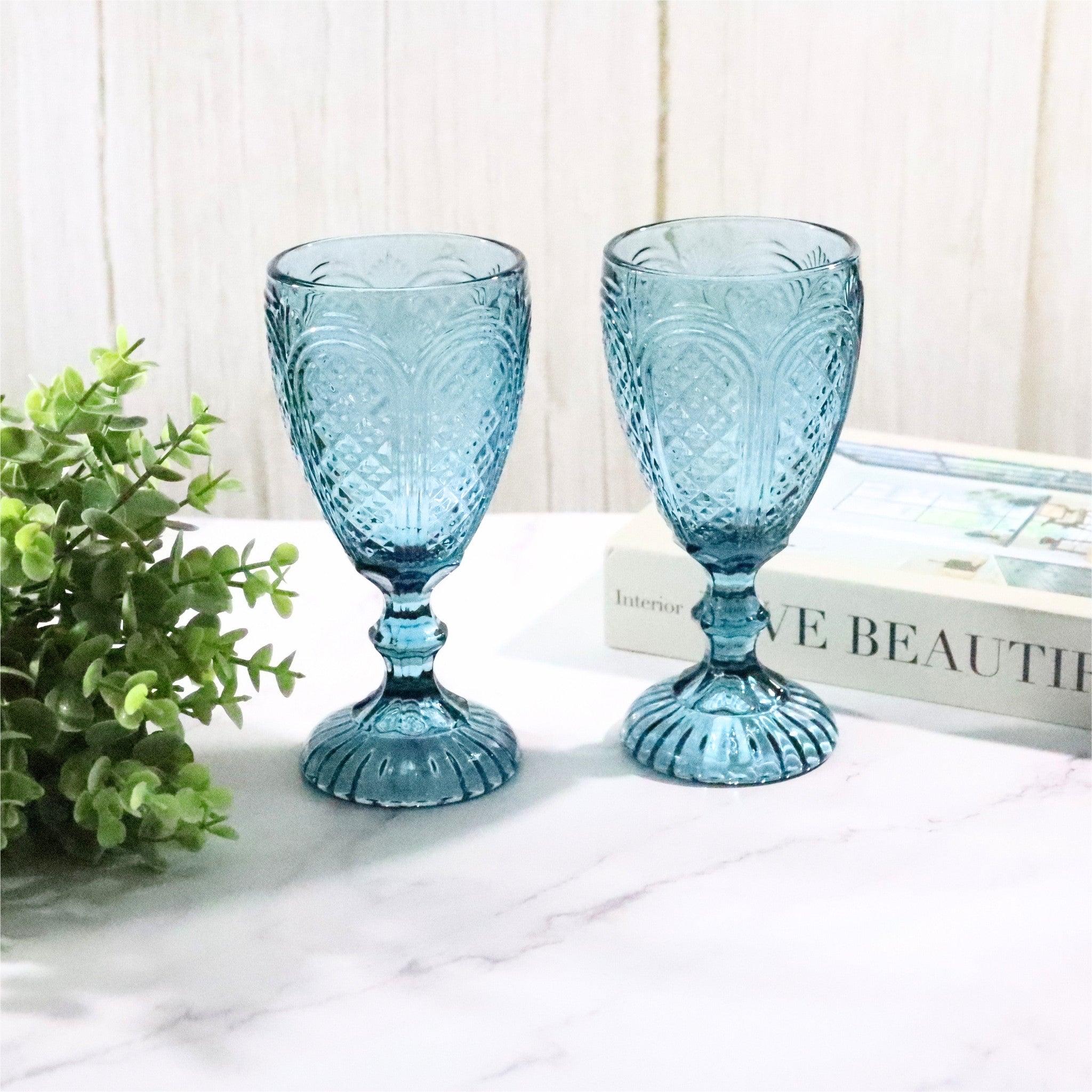 Two blue textured glass goblets on a white surface with a plant and book in the background.