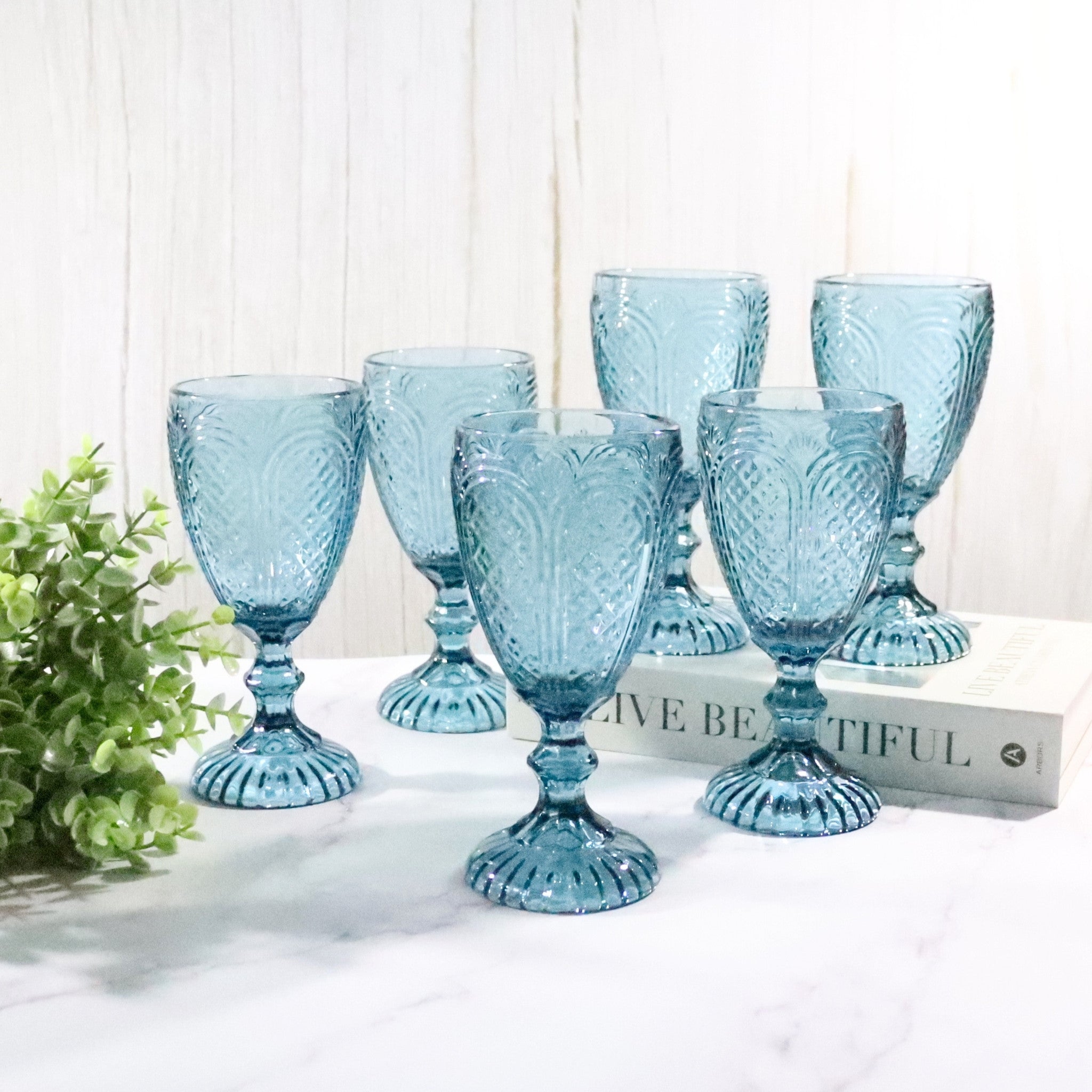 Set of blue glass goblets on a white surface with a plant and book in the background.