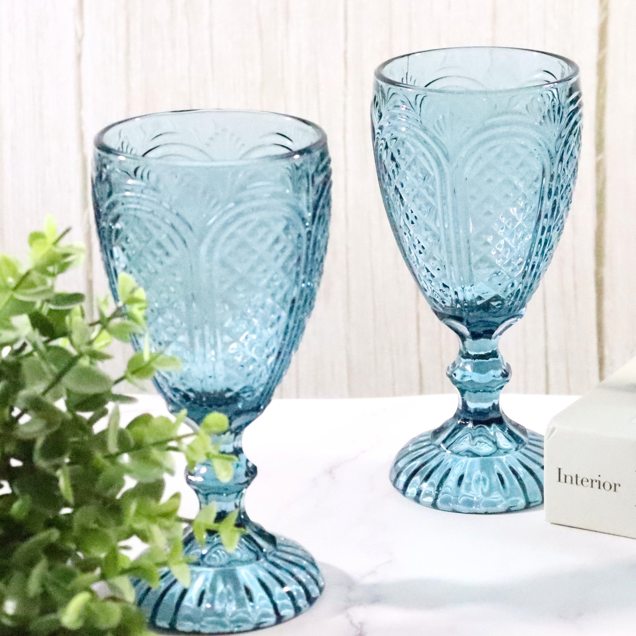 Two blue textured glass goblets on a white surface with a plant in the foreground.