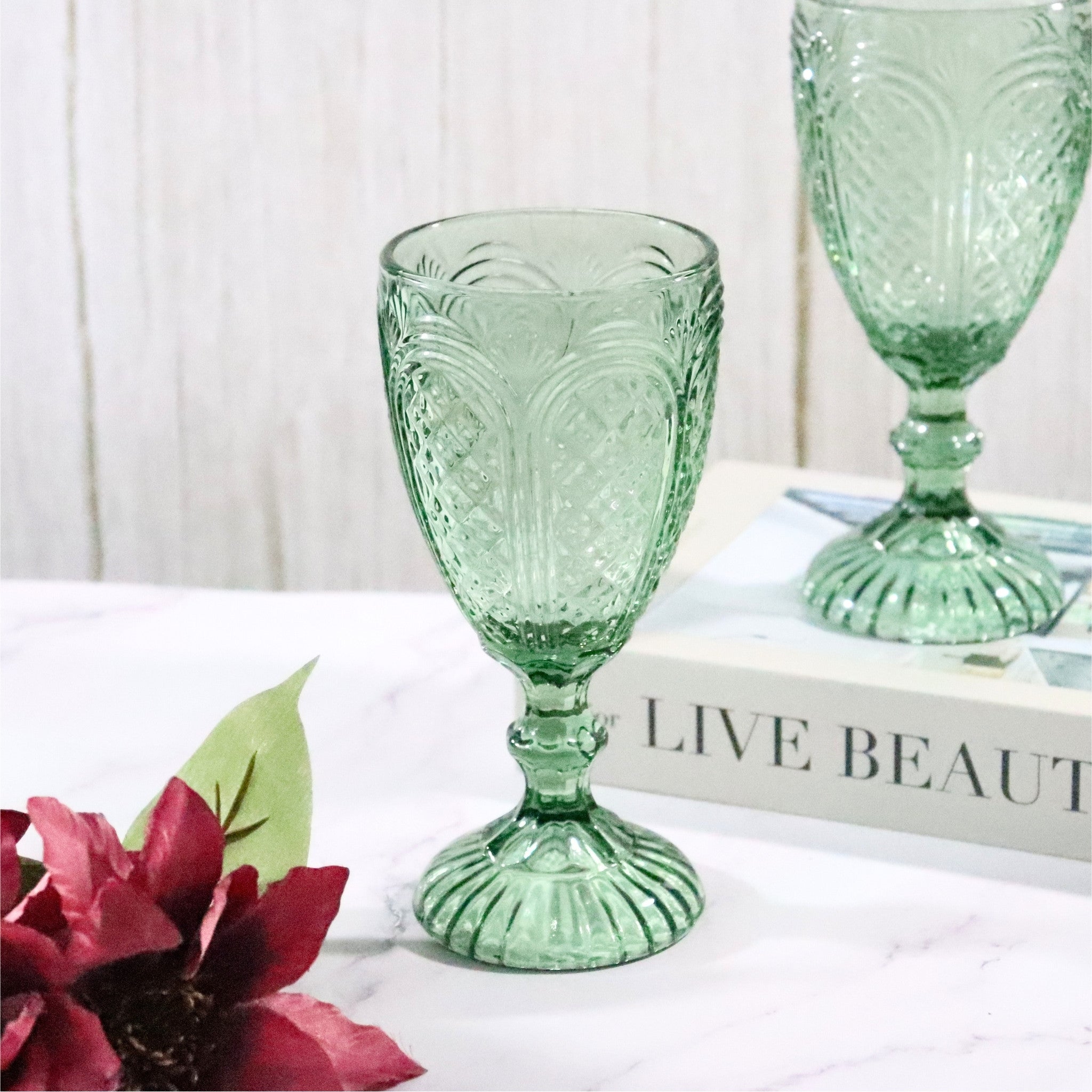 Two green glass goblets on a white surface with a book and red flowers in the background.