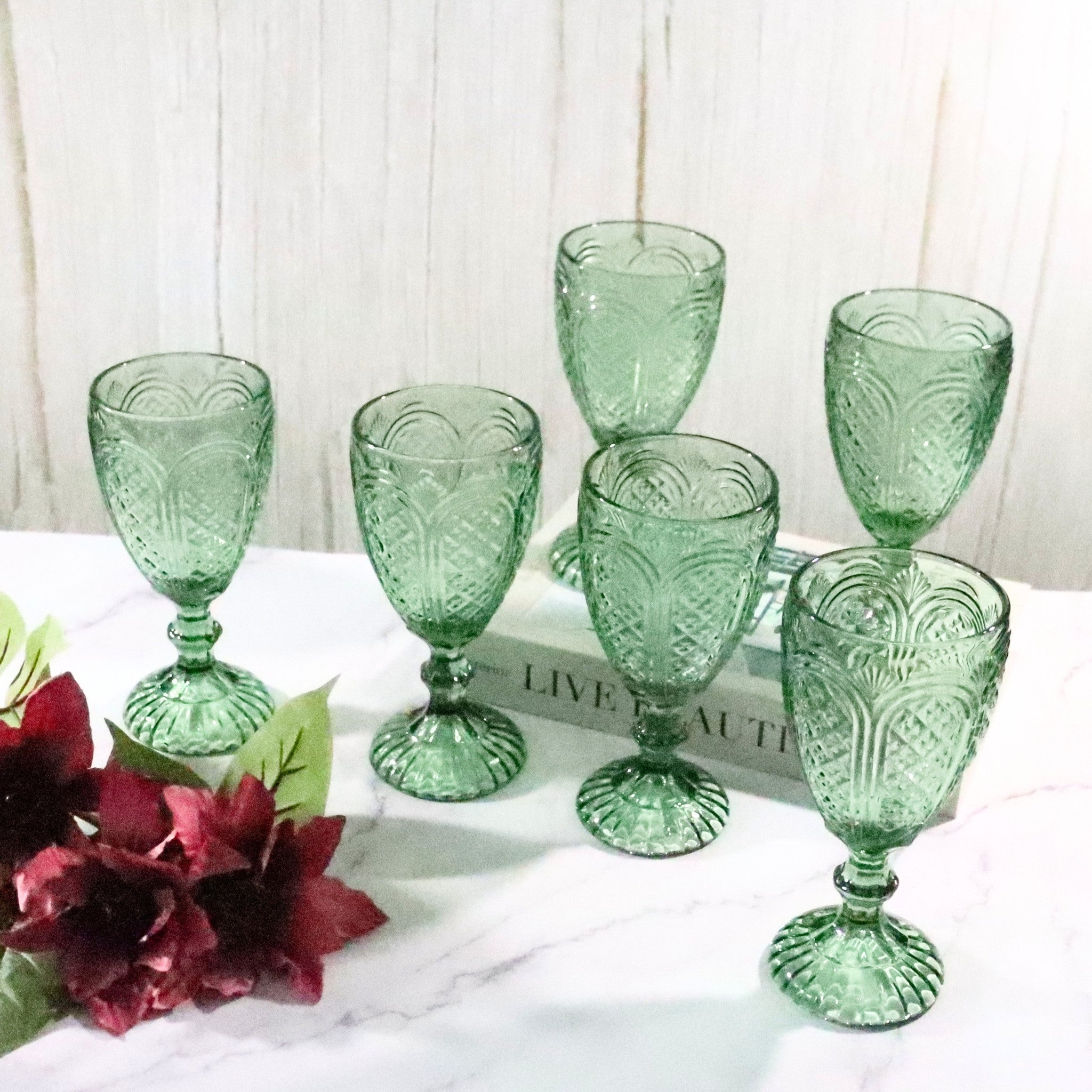 Set of six green glass goblets on a white surface with flowers and books in the background.