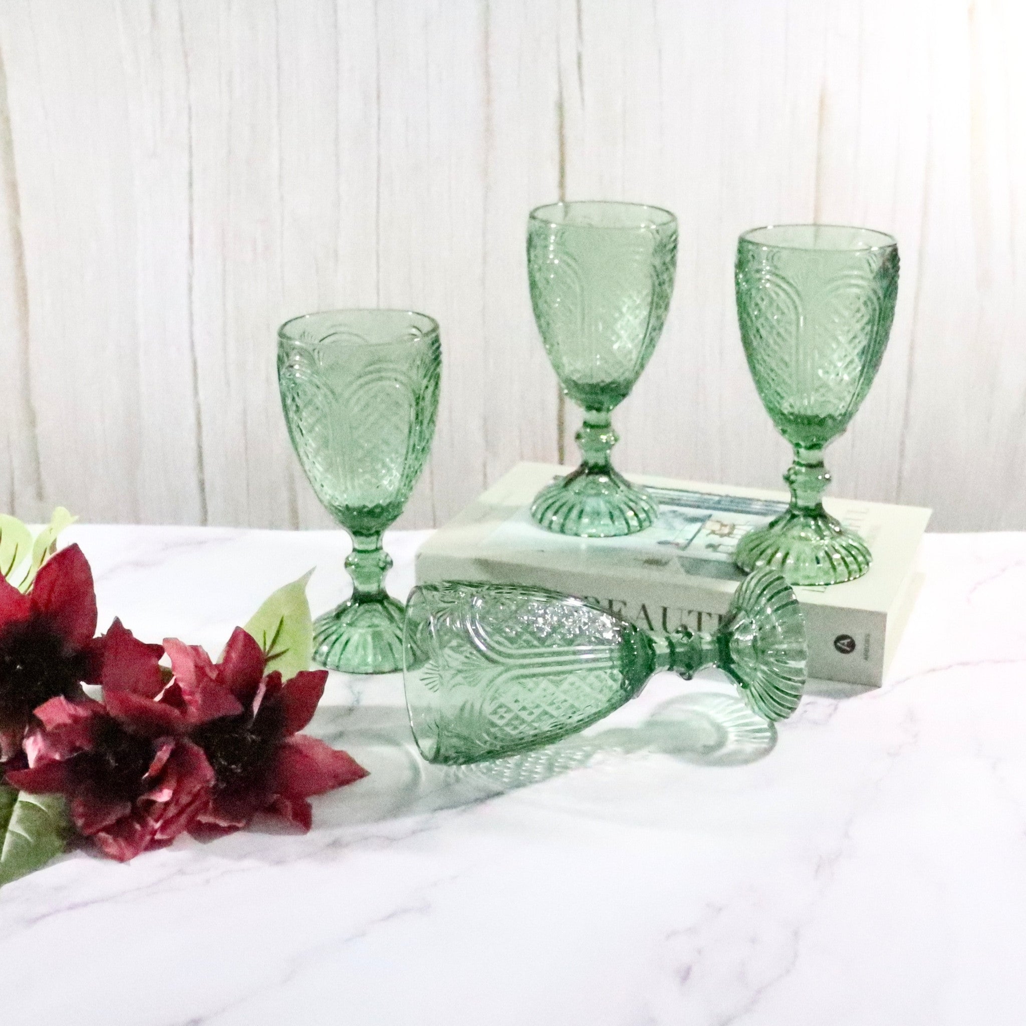 Green glass goblets on a white surface with red flowers and books in the background