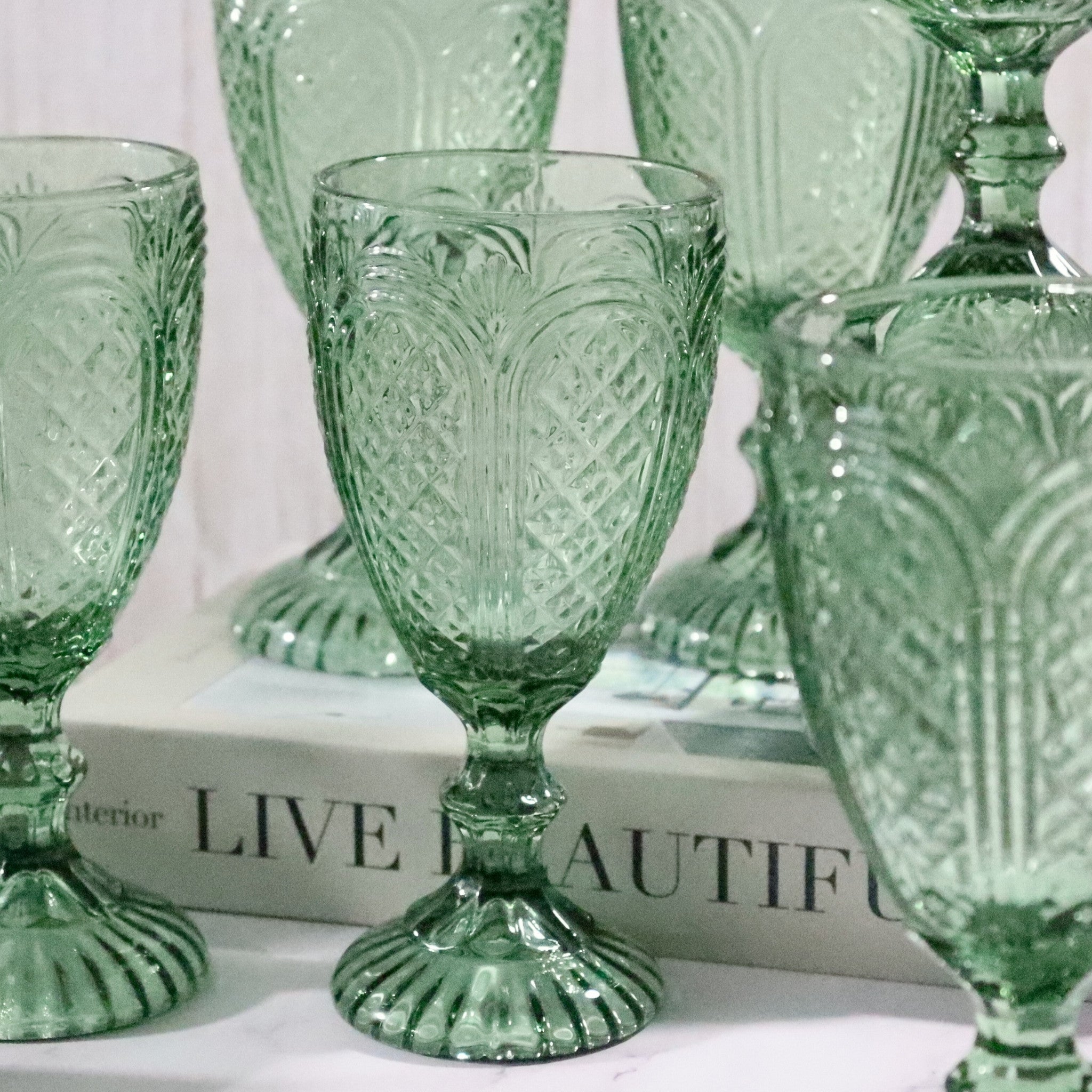 Set of green patterned glass goblets on a white surface with a blurred book in the background.