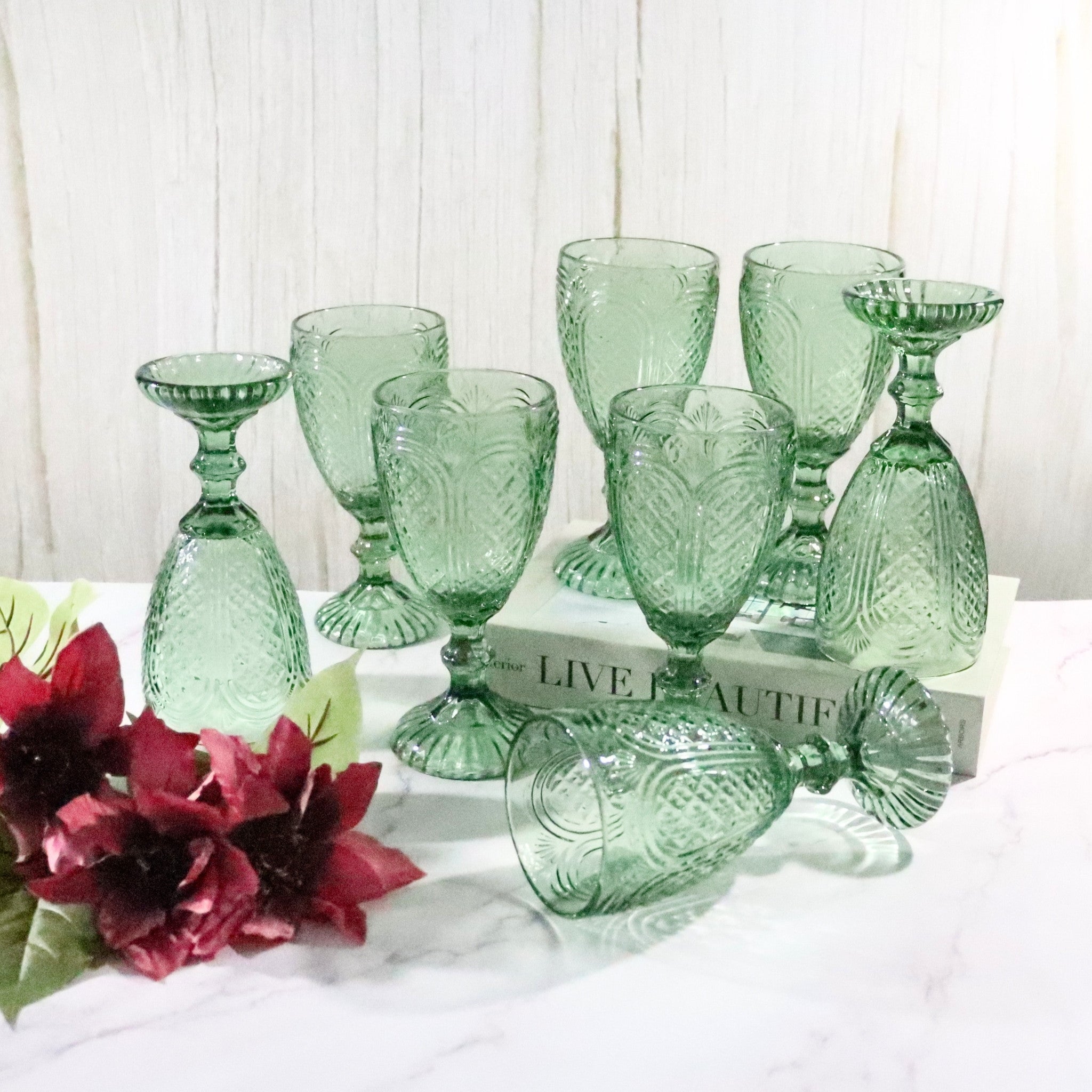 Set of green glass goblets on a white surface with red flowers and a book in the background.