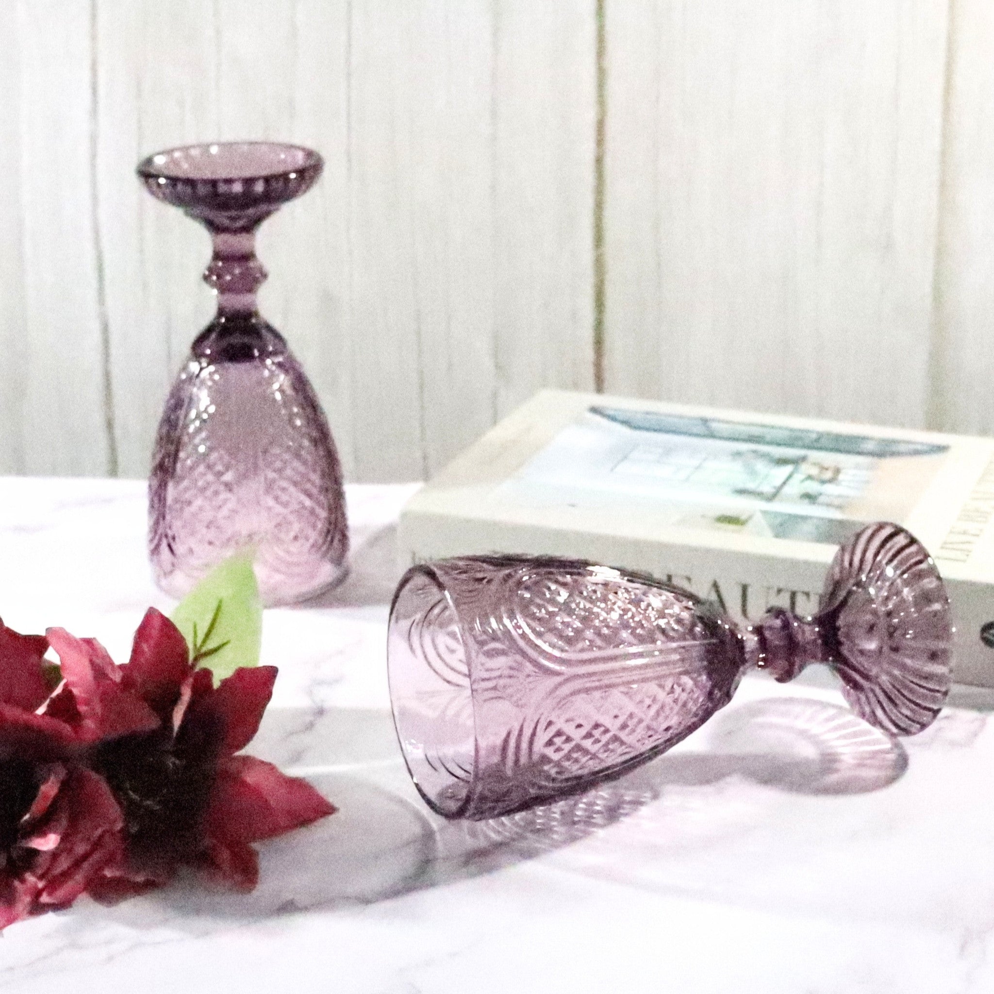 Purple glass goblet and candle holder on a white surface with flowers and books in the background