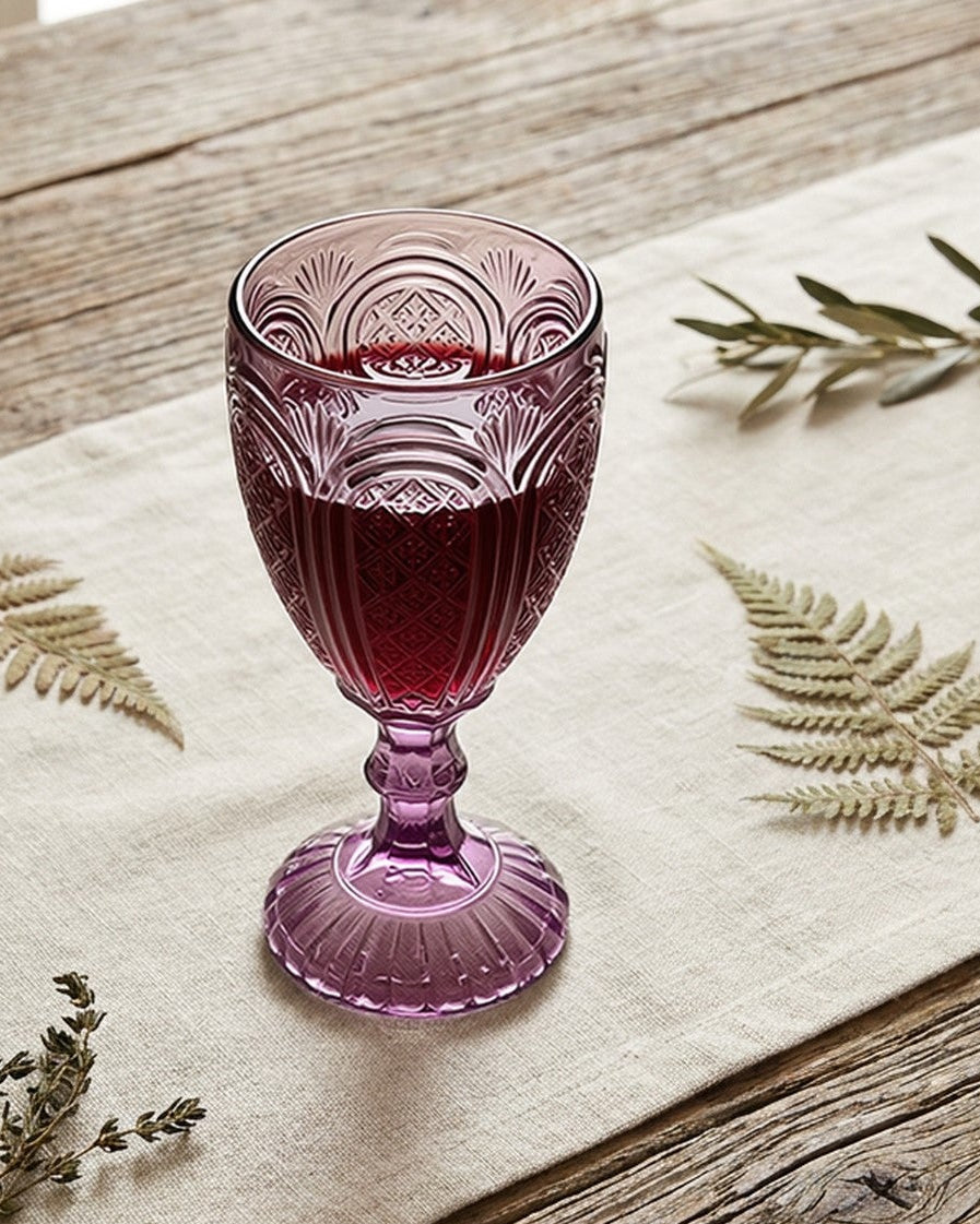 Purple glass goblet and candle holder on a white surface with flowers and books in the background