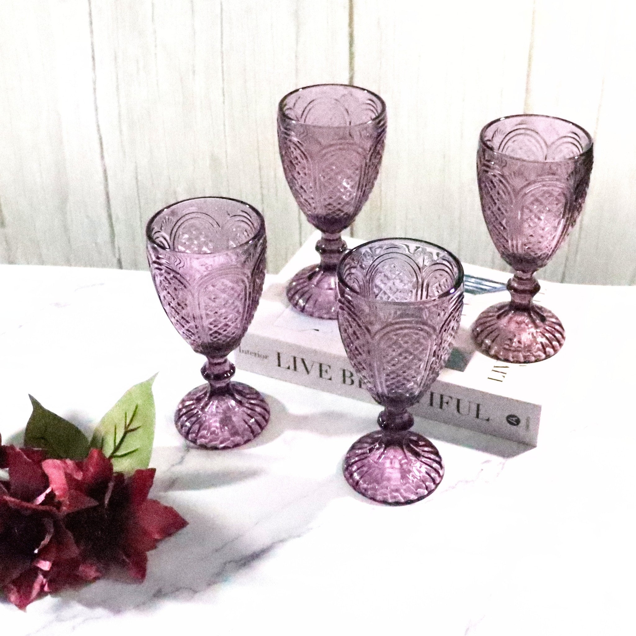 Set of five purple glass goblets on a white surface with a floral arrangement.