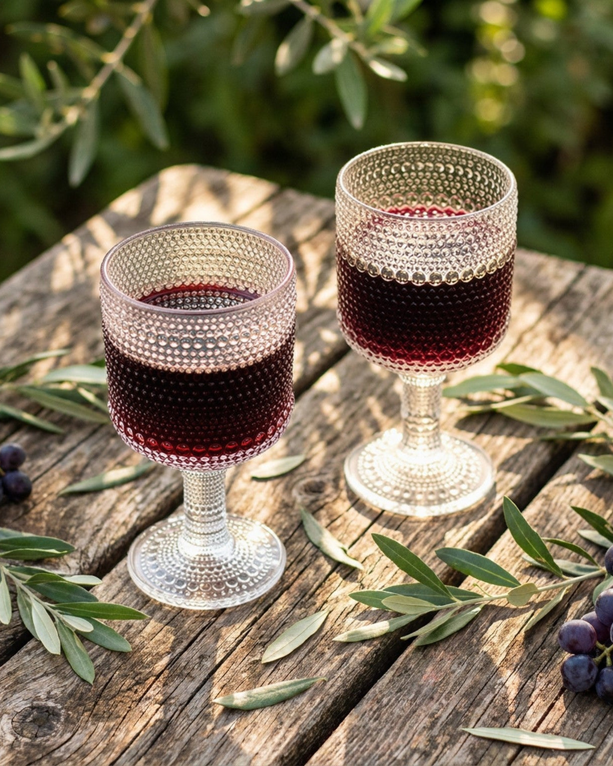 Two glasses of red wine on a wooden table with grapes and olive branches.