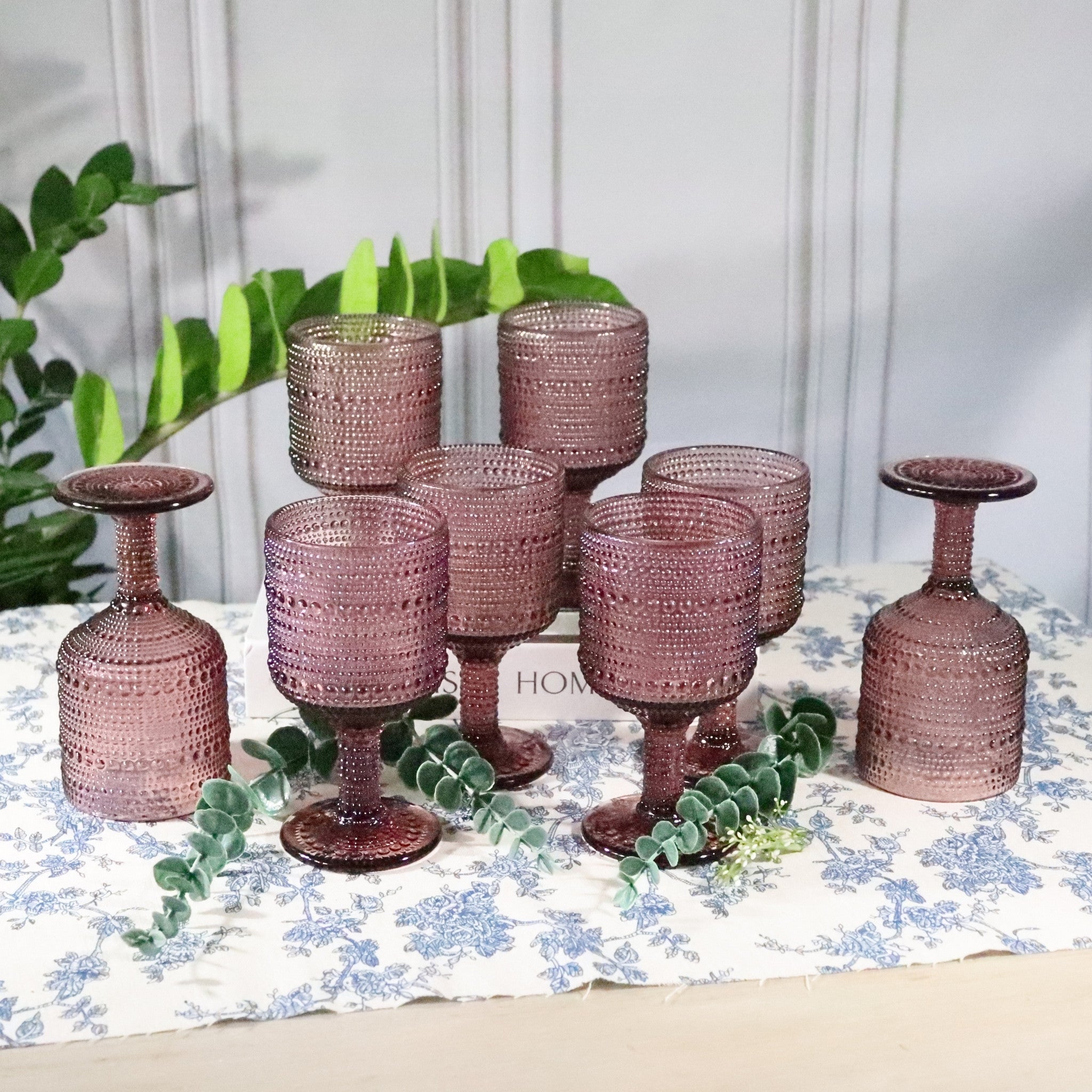 Set of purple hobnail bead glass goblets on a floral tablecloth with greenery in the background