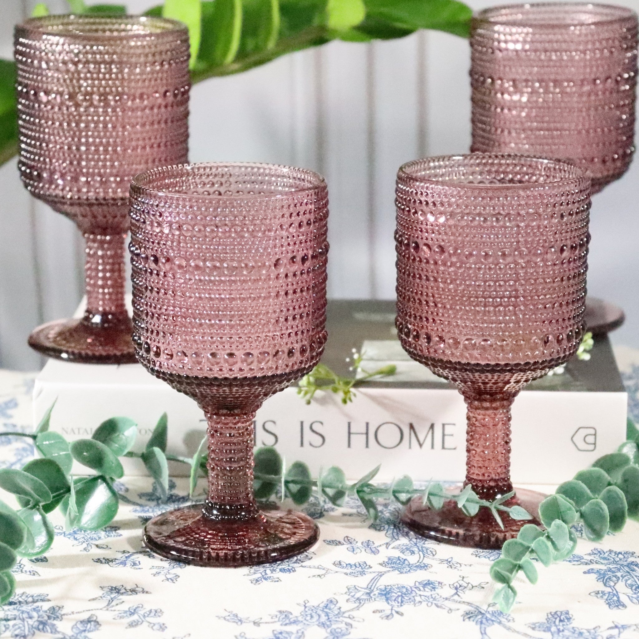 Set of four pink textured glass goblets on a decorative table with greenery and 'This is Home' sign.