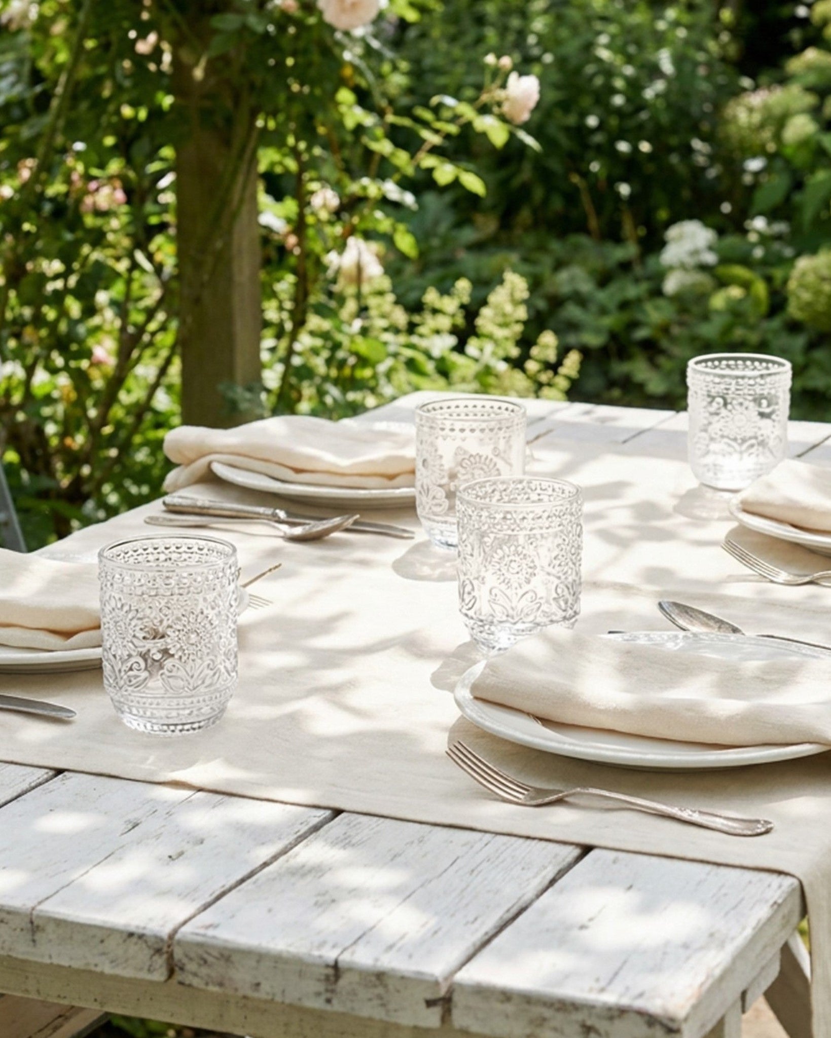 Dining table set with plates, glasses, and cutlery on a wooden table outdoors with greenery in the background.
