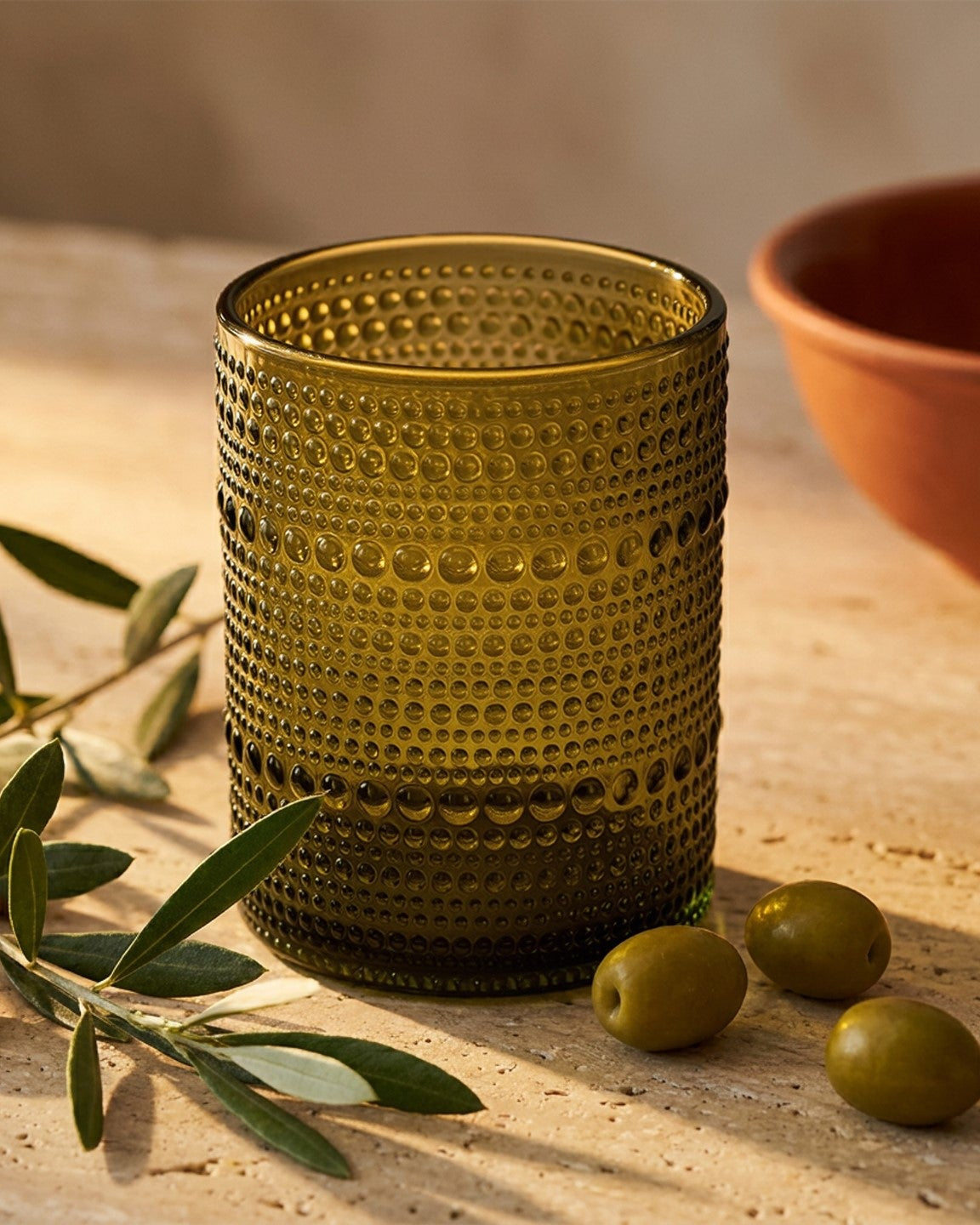 Textured brown glass candle holder on a wooden surface with olives and an olive branch.