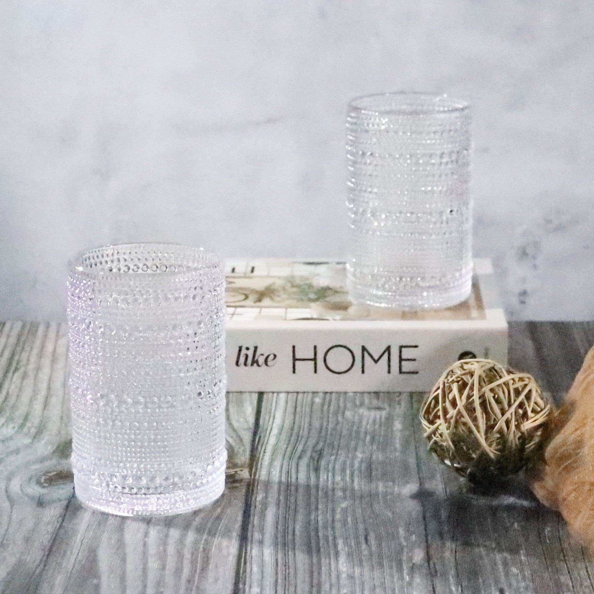 Two beaded hobnail clear drinking glasses on a wooden surface with 'like HOME' box in the background.
