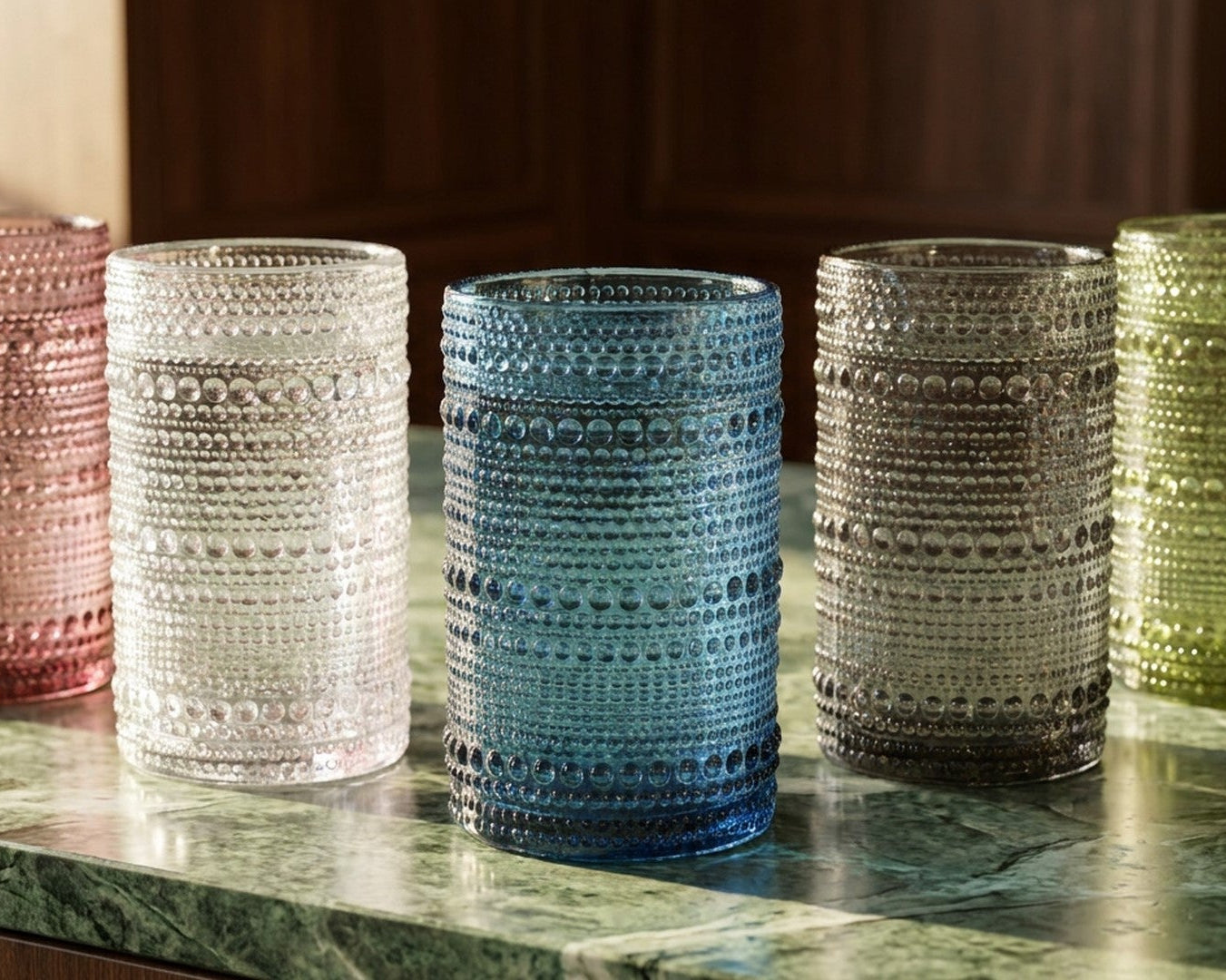 Two green textured glass tumblers on a wooden surface with a book and flower in the background.