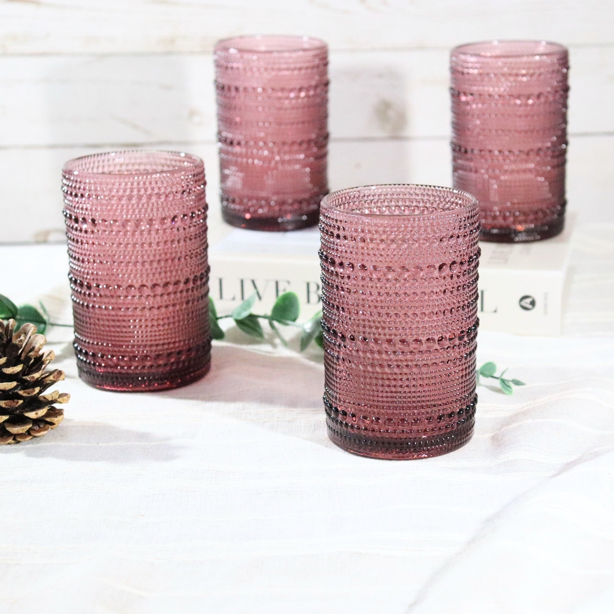 Set of four pink textured glass tumblers on a white surface with greenery and a pine cone.